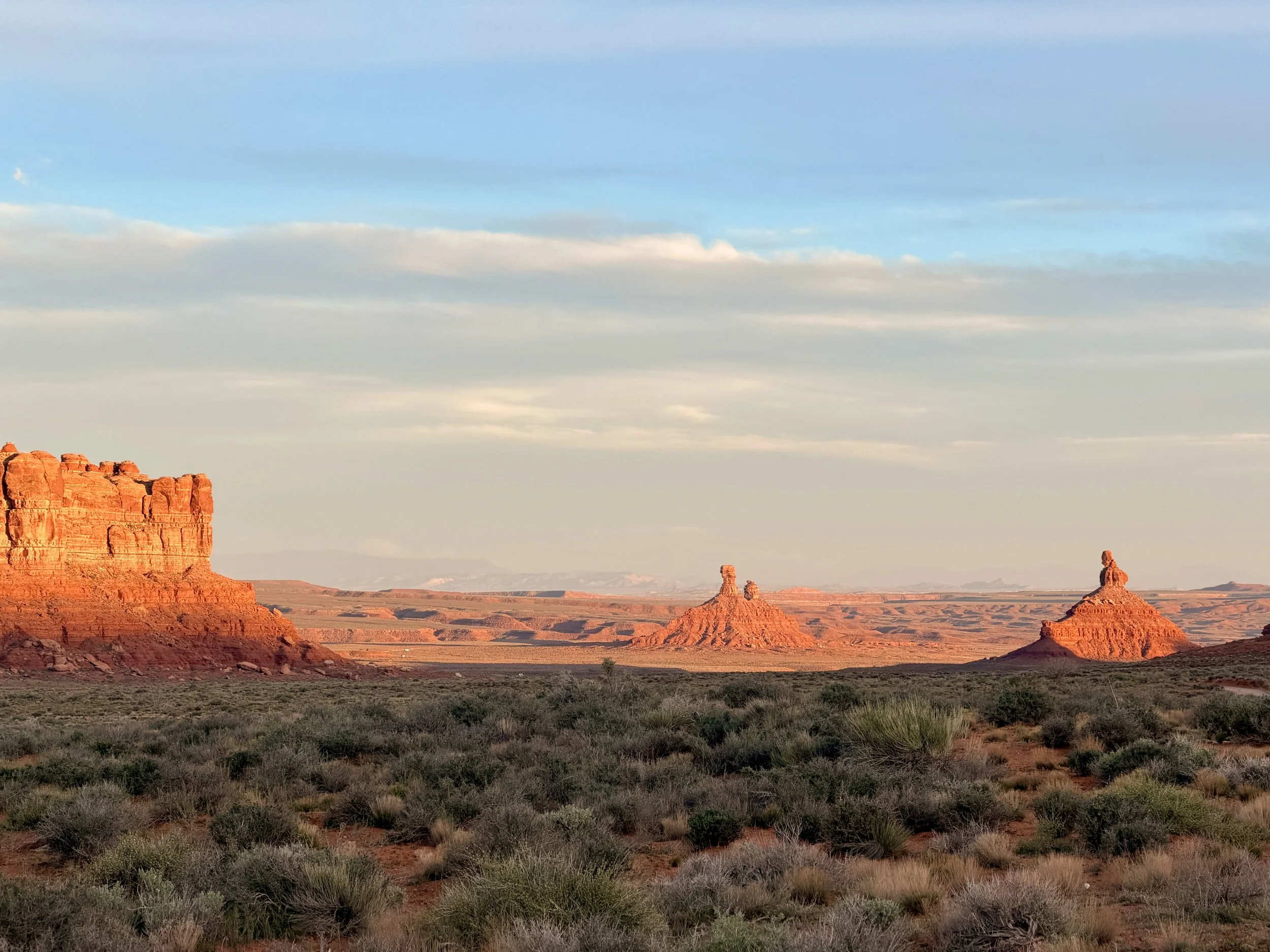 A desert landscape with red rock formations in the foreground and middle ground, green shrubs across the terrain, and a cloudy sky overhead.
