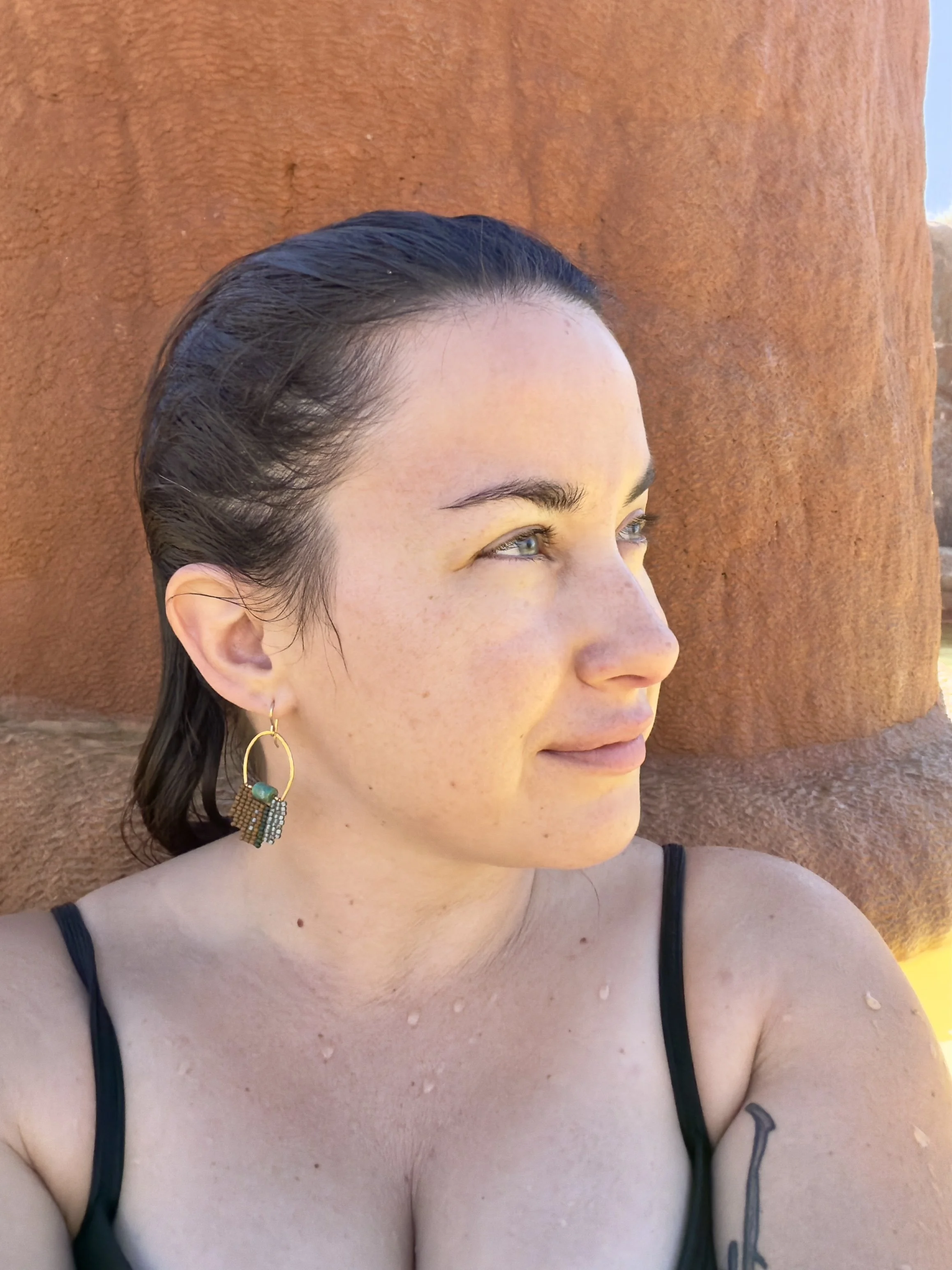 A woman with dark hair tied back, wearing dangling earrings with beads, in front of a textured orange-brown backdrop at a hot springs.