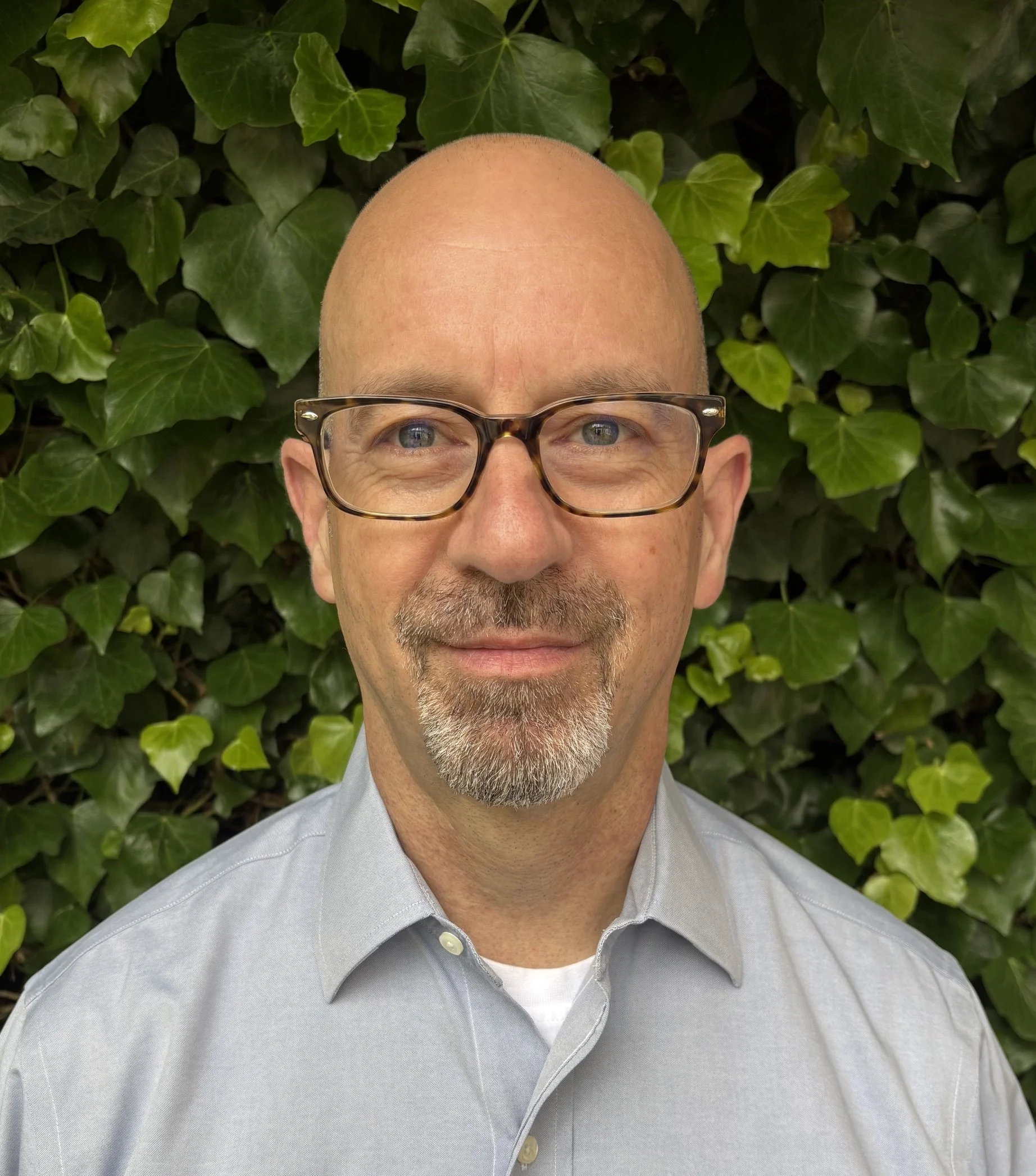 A close-up of a bald middle-aged man with glasses standing in front of green ivy leaves.