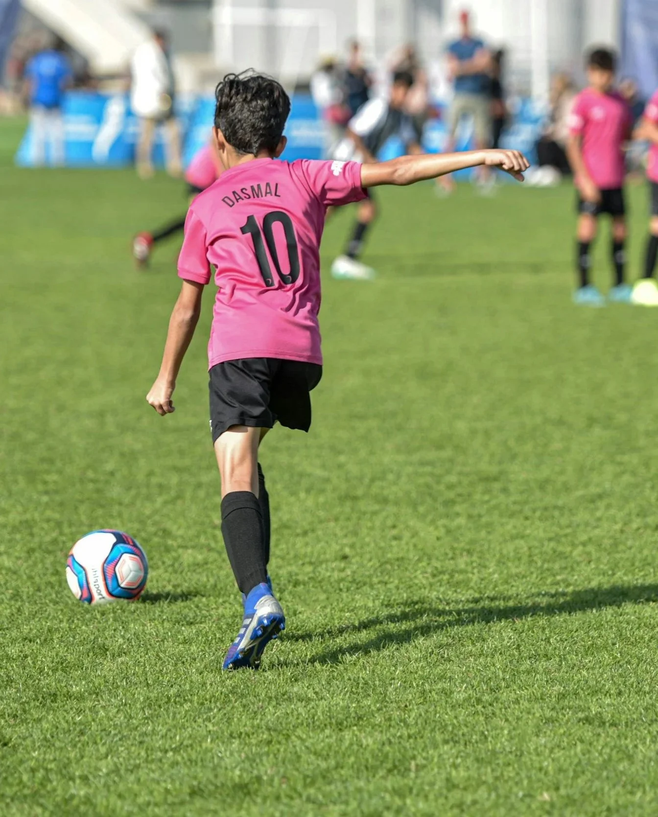A young soccer player in a pink jersey with number 10 and the name Dasmal dribbling a soccer ball on a green field, with other players and spectators in the background.