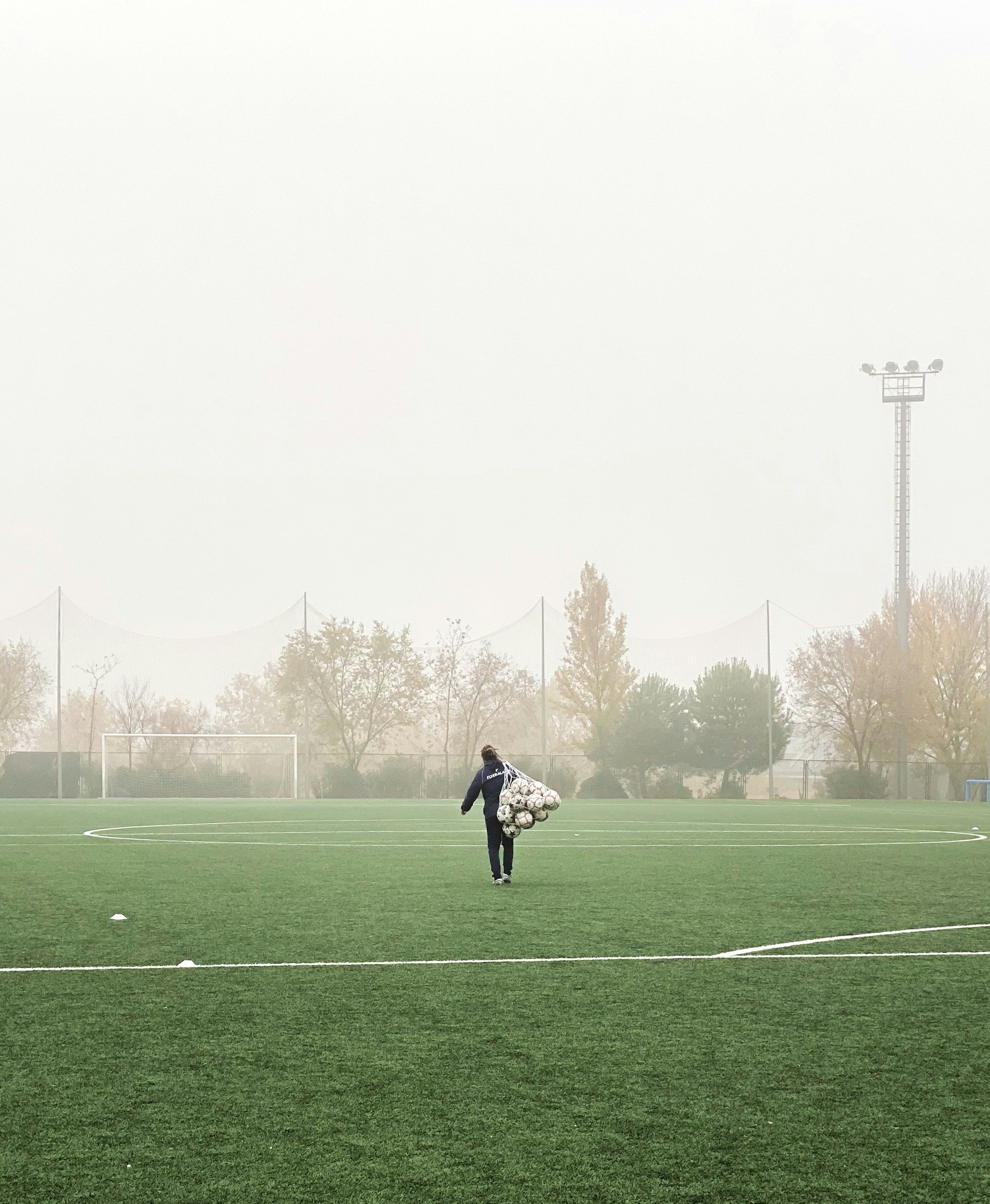 Person walking on a soccer field carrying a large bundle of soccer balls, with goal and trees in the background, foggy sky.