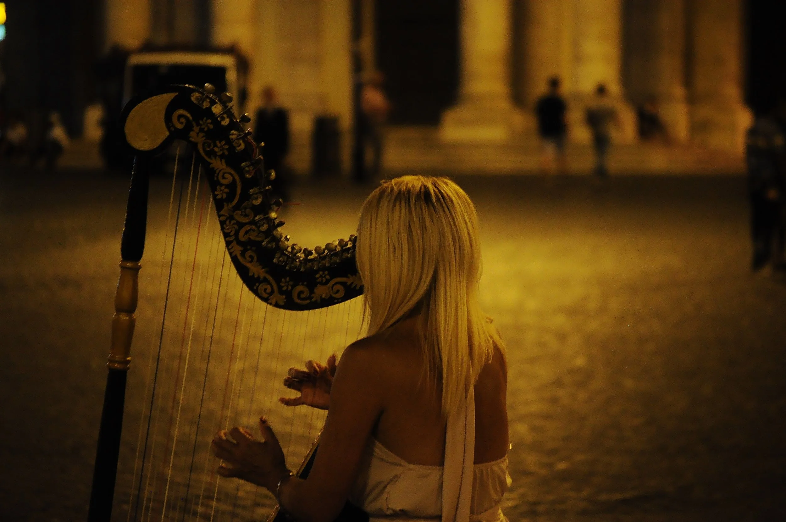A woman with long blonde hair playing a harp outdoors at night, with a large illuminated building in the background.