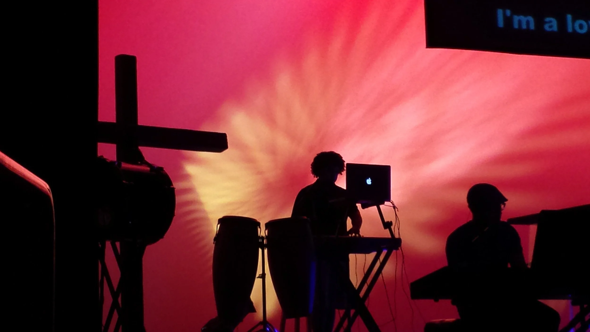 Silhouetted musicians performing on stage against a vivid pink and orange sunset background, with electronic music equipment and a person using a laptop.