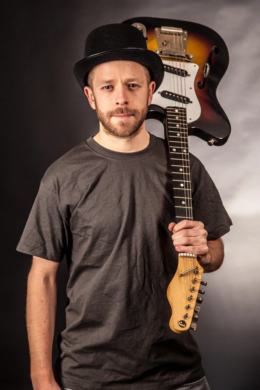 A man wearing a black hat and gray t-shirt, holding an electric guitar over his shoulder, standing against a dark background.