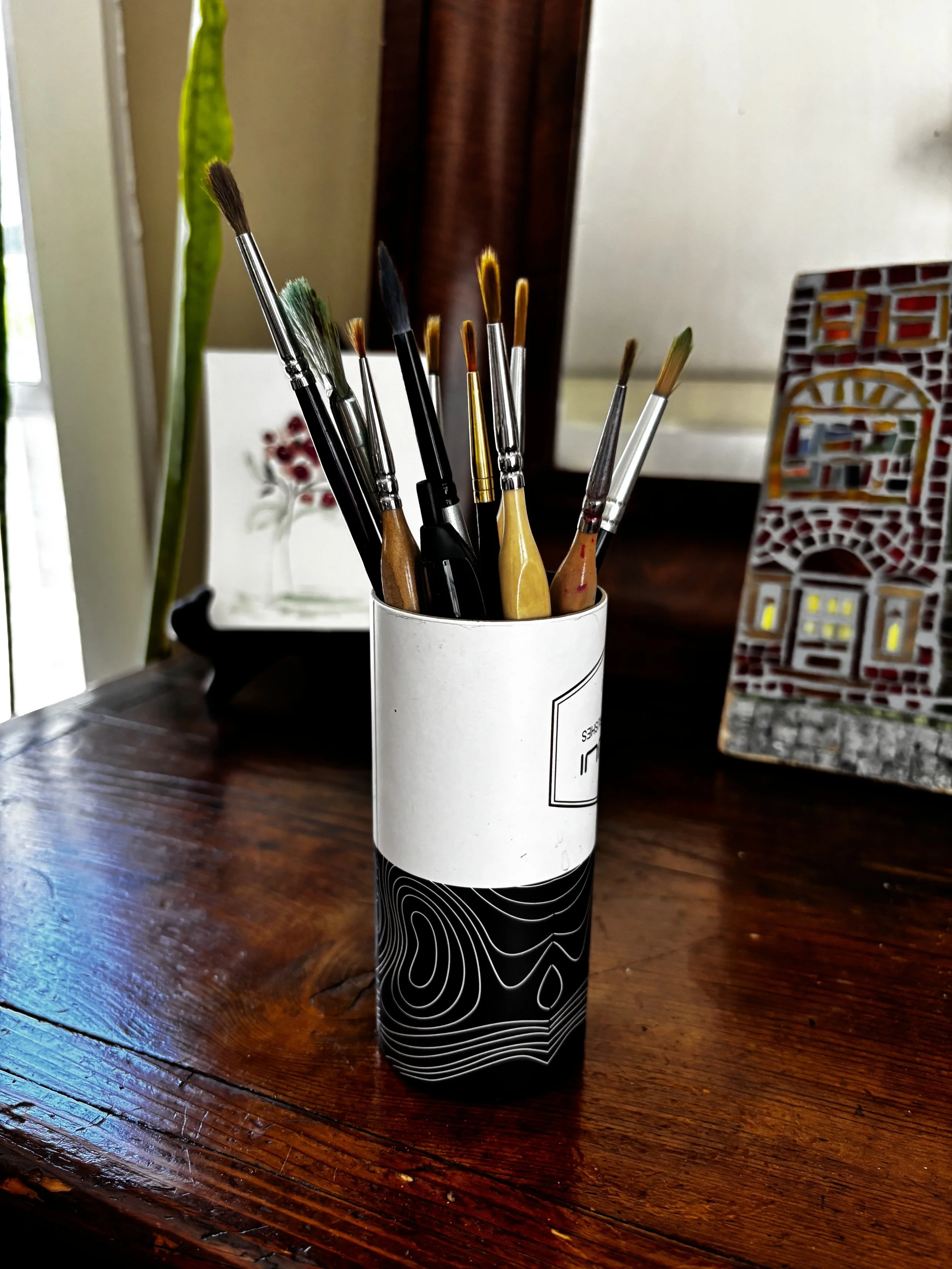A white container with black abstract line art design holds various paintbrushes on a wooden table. In the background, there is a green plant, a decorative tile, and a framed picture with a floral design.