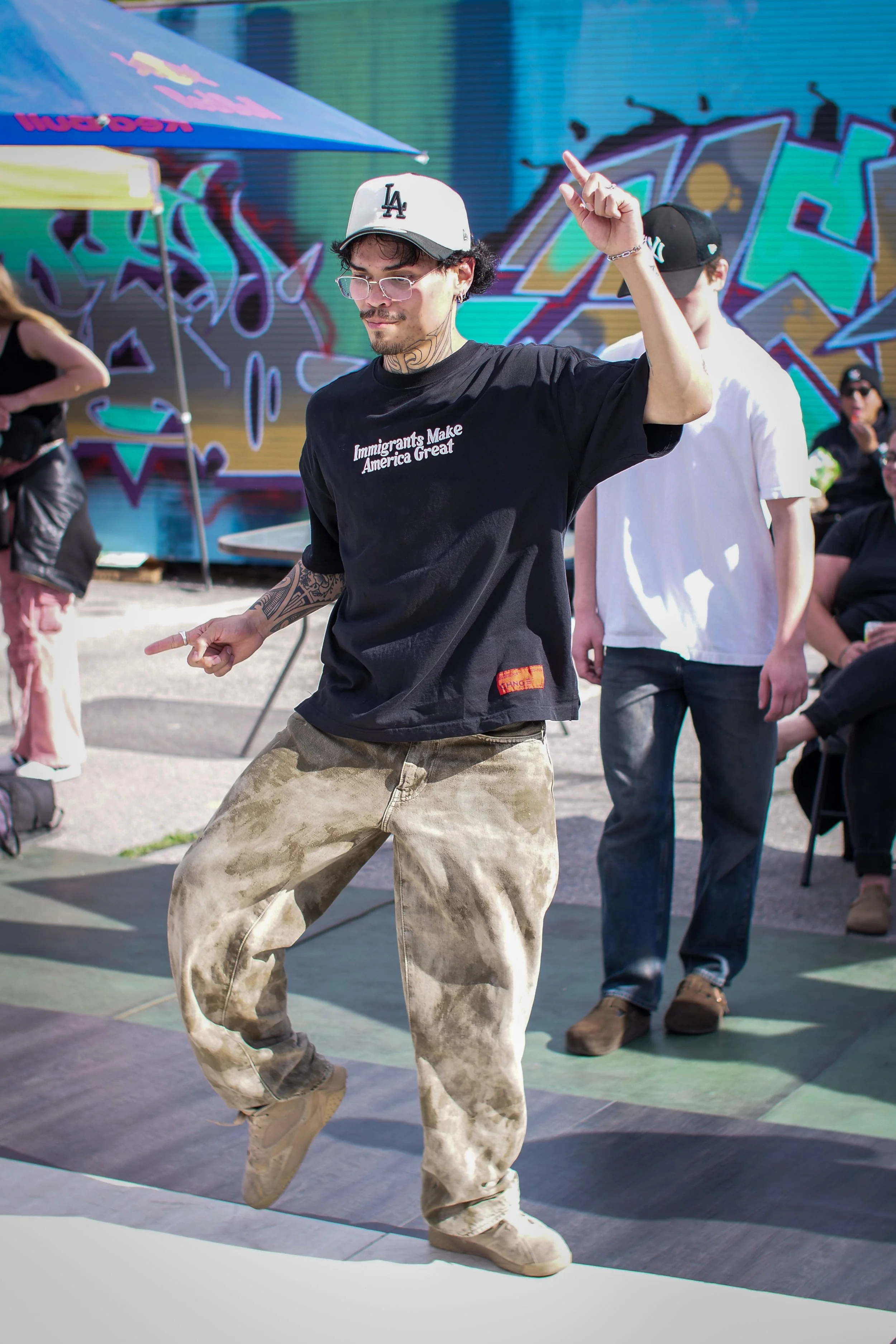 Young man dancing outdoors in front of colorful graffiti, wearing a LA baseball cap, glasses, black t-shirt with the text "Immigrants Make America Great," and loose camera-matched camouflage pants.