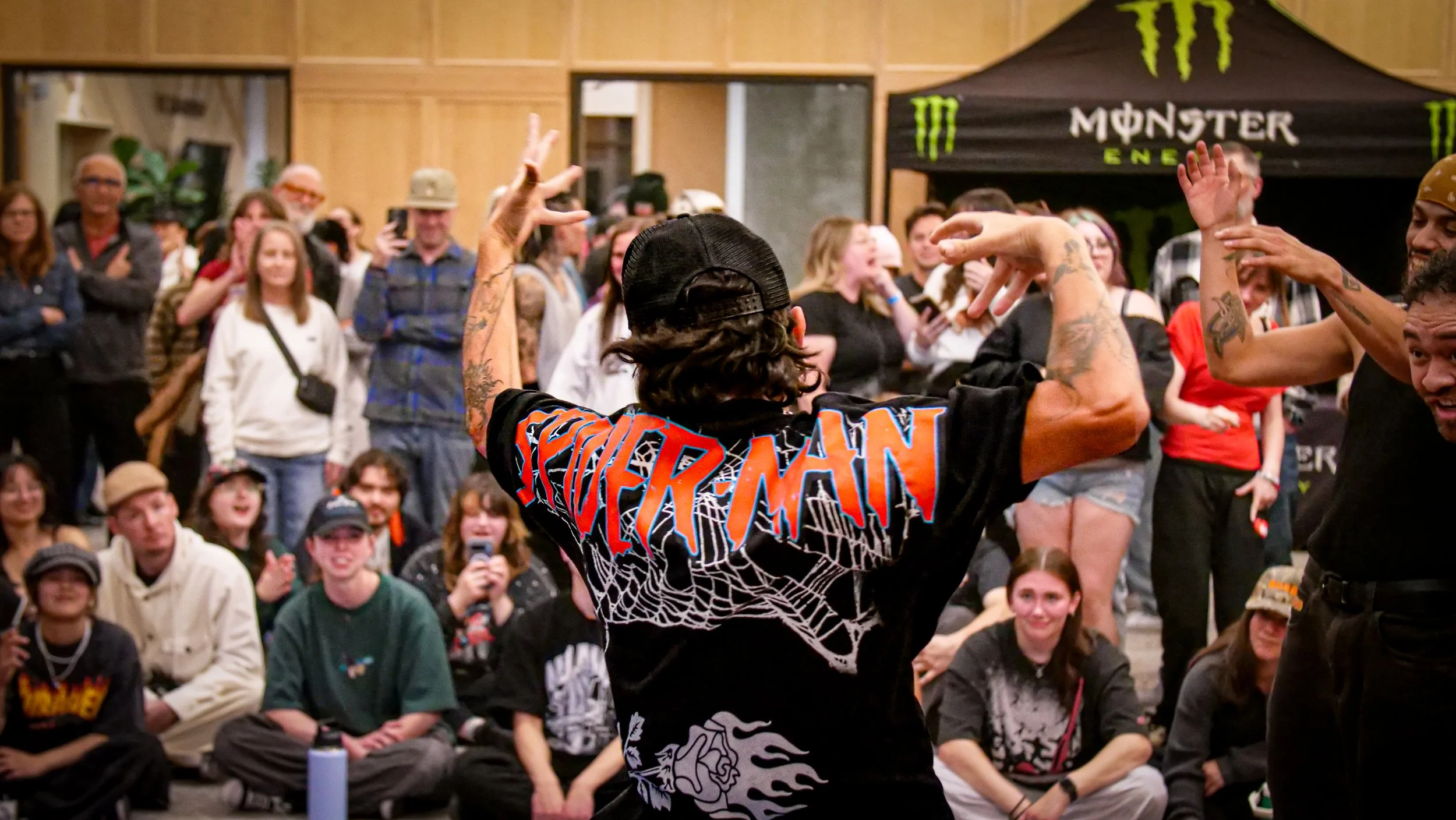 A performer with tattoos and dark curly hair, wearing a black cap and a black hoodie with colorful text, is dancing or performing in front of an audience at an indoor event with a Monster Energy tent in the background.