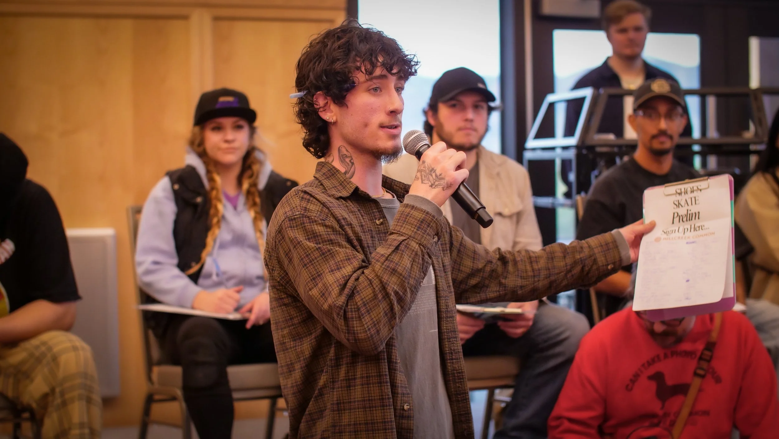 A young man with curly hair and tattoos on his neck is speaking into a microphone during a meeting or discussion, with several other people seated around him.