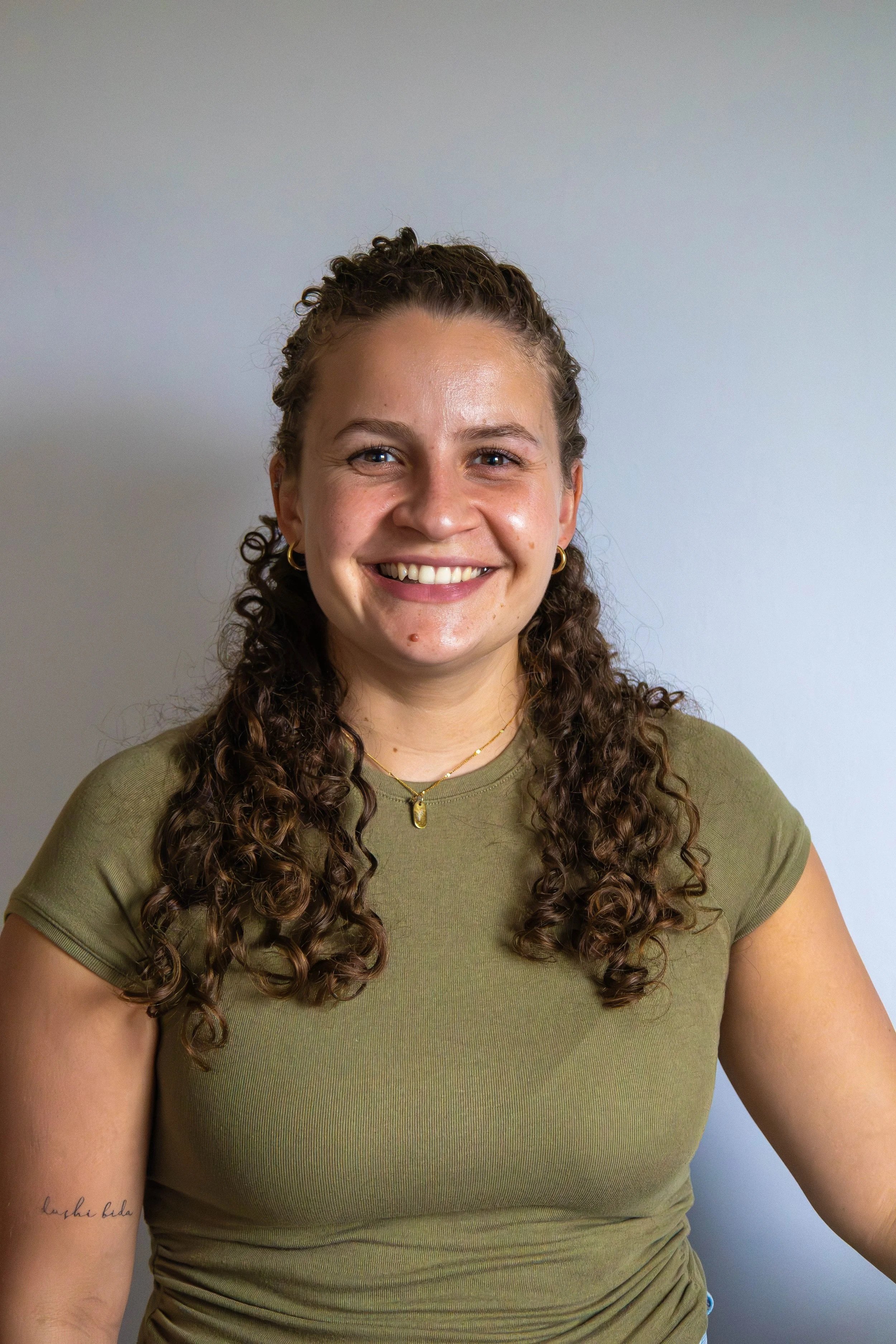 Smiling woman with curly hair wearing a green t-shirt and gold jewelry, standing against a plain background.