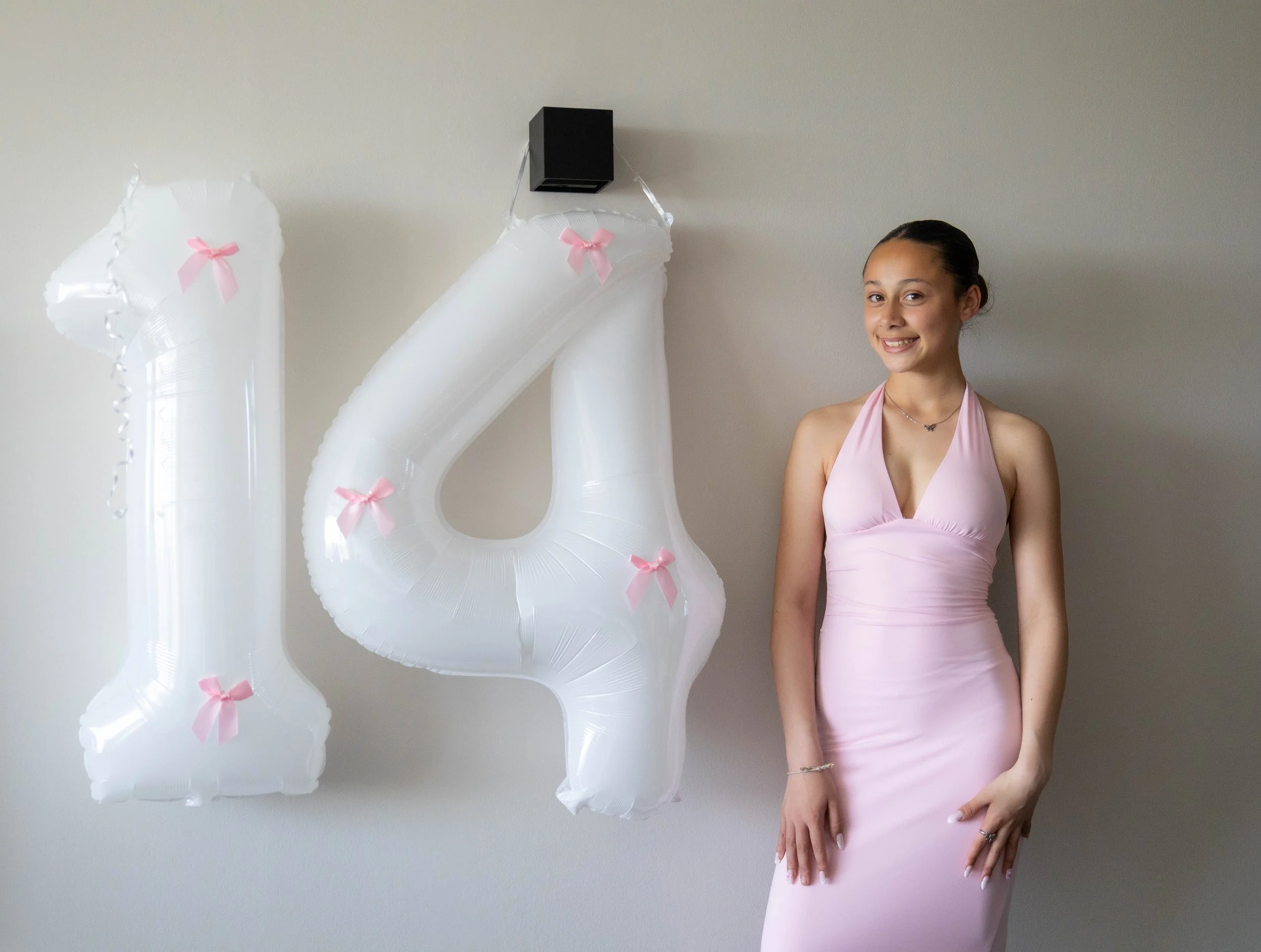 A young woman in a pink dress smiling while standing beside white balloons shaped like the number four, decorated with small pink bows, against a plain wall.