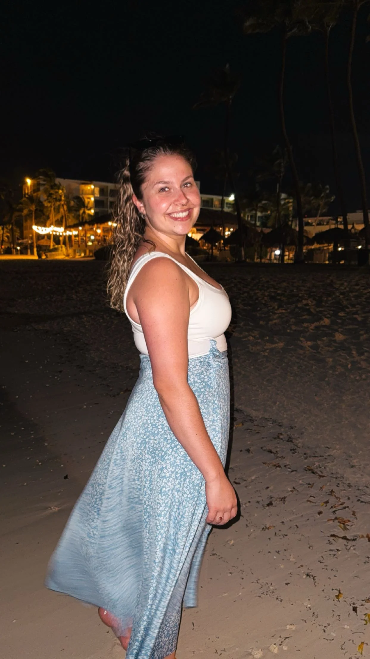 A woman standing on a beach at night, smiling, wearing a white tank top and a flowing light blue skirt, with palm trees and hotel buildings illuminated in the background.