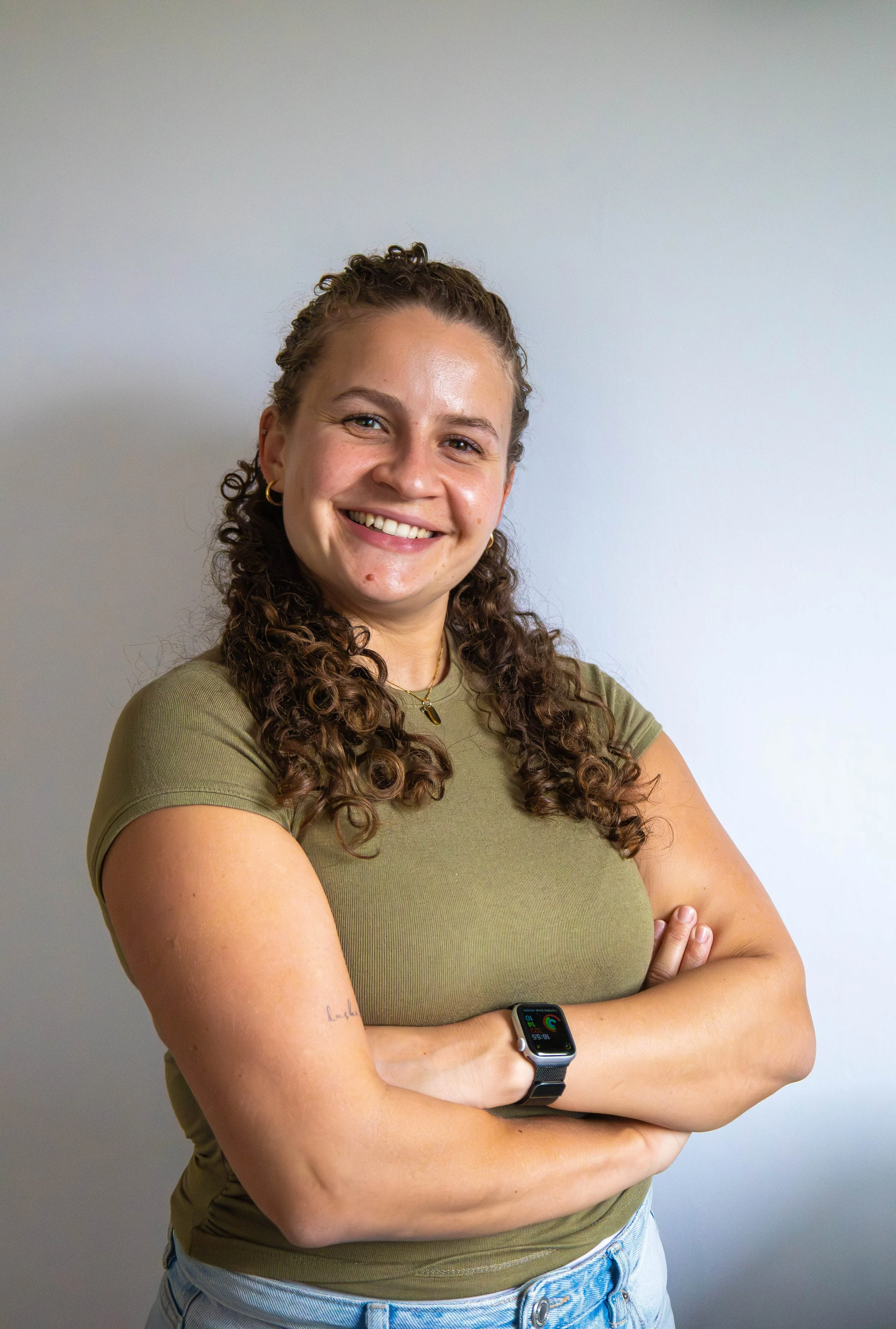 A young woman with curly hair smiling, arms crossed, wearing a green t-shirt and a smartwatch, standing against a plain light-colored wall.