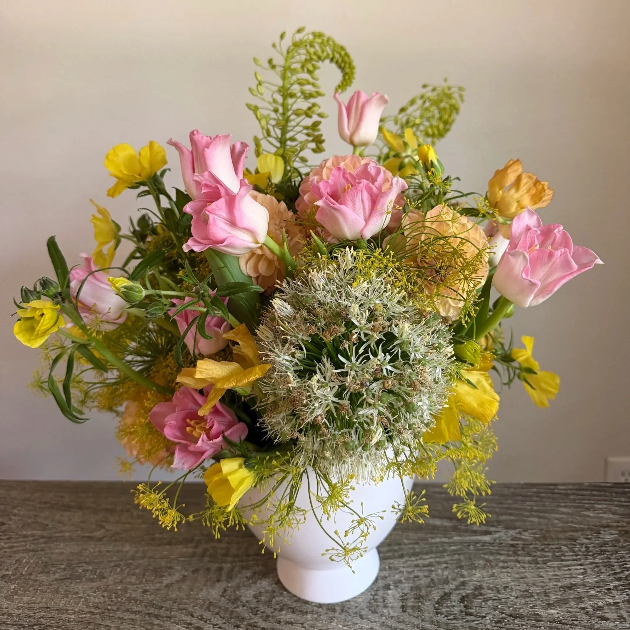 A bouquet of pink, yellow, and white flowers in a white vase on a wooden surface.