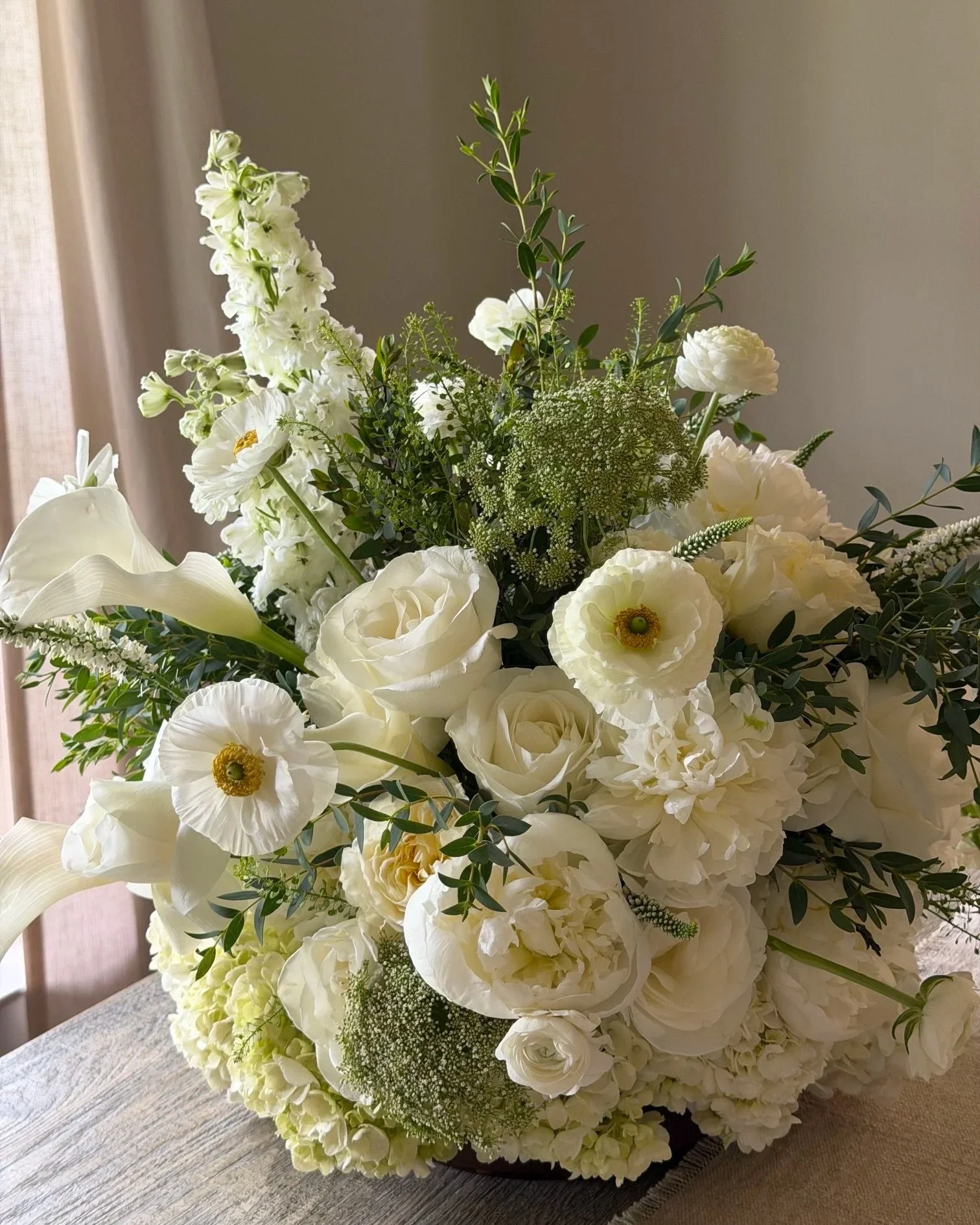 A large floral arrangement with white flowers and green foliage in a dark container on a wooden table near a window.