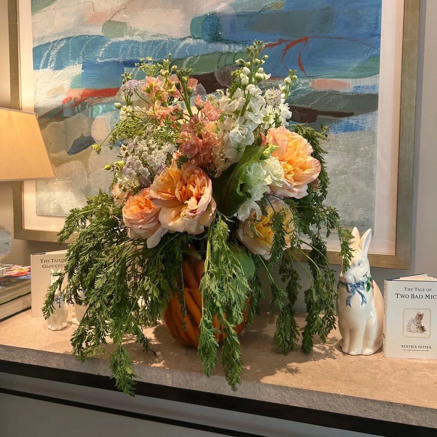 A bouquet of flowers in an orange vase, featuring white, pink, and peach blossoms with green foliage, placed on a table with books and decorative items including a ceramic rabbit, with an abstract painting in the background.