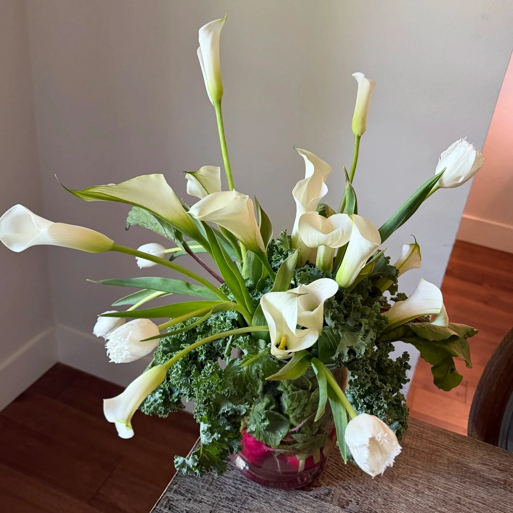 A bouquet of white calla lilies, pink flowers, and green foliage in a glass vase on a wooden table.