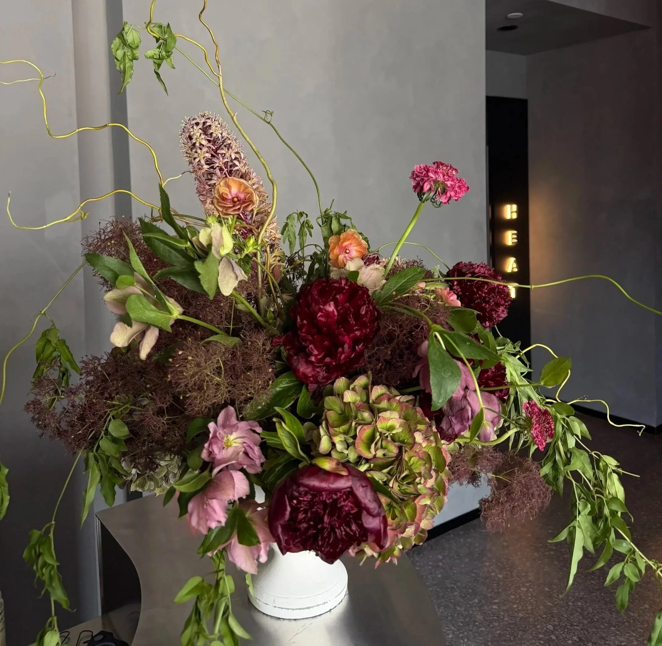 A large floral arrangement of pink, burgundy, and purple flowers with green leaves sits in a white vase on a dark countertop against a gray wall, with illuminated signs in the background.