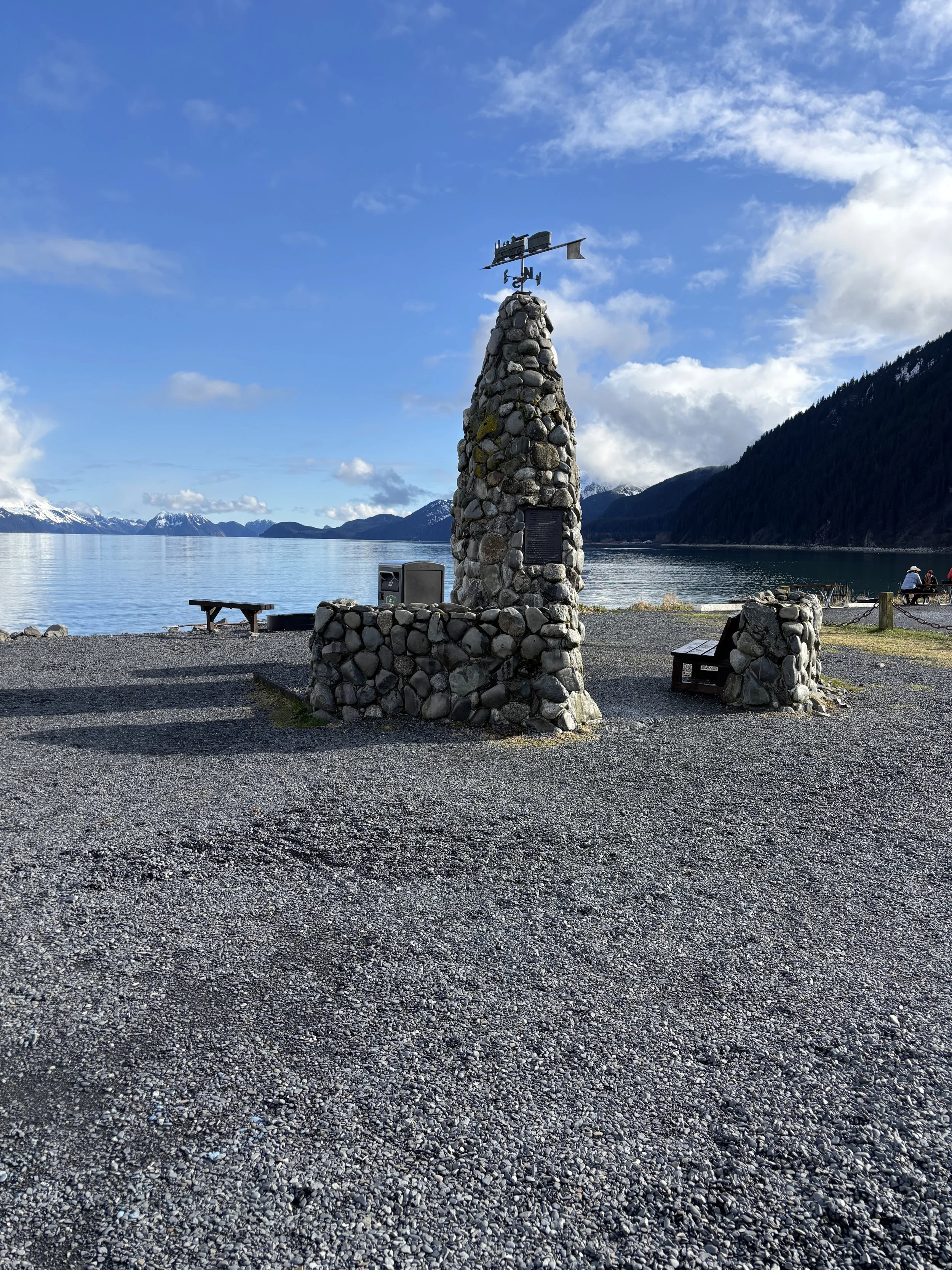 A stone monument with a weather vane on top by a lakeside, with mountains and partly cloudy sky in the background.