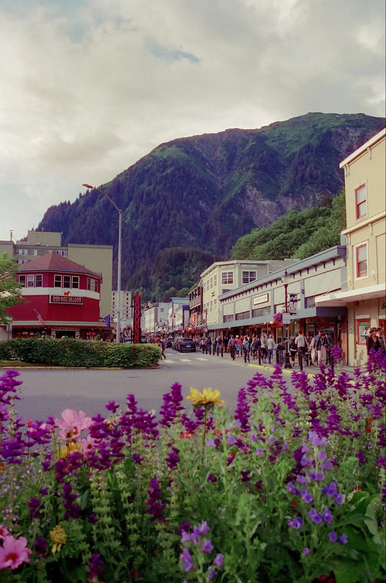 A lively street scene with people walking on the sidewalk, colorful flowers in the foreground, and a mountain in the background.