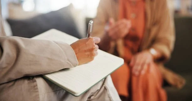 A person sitting on a couch writing in a notepad with a pen, while another person sits nearby.