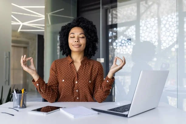 A woman with curly hair sitting at a desk with her eyes closed, practicing meditation or mindfulness with her hands in a mudra gesture. There is a laptop, smartphone, and office supplies on the desk, and a glass window in the background.