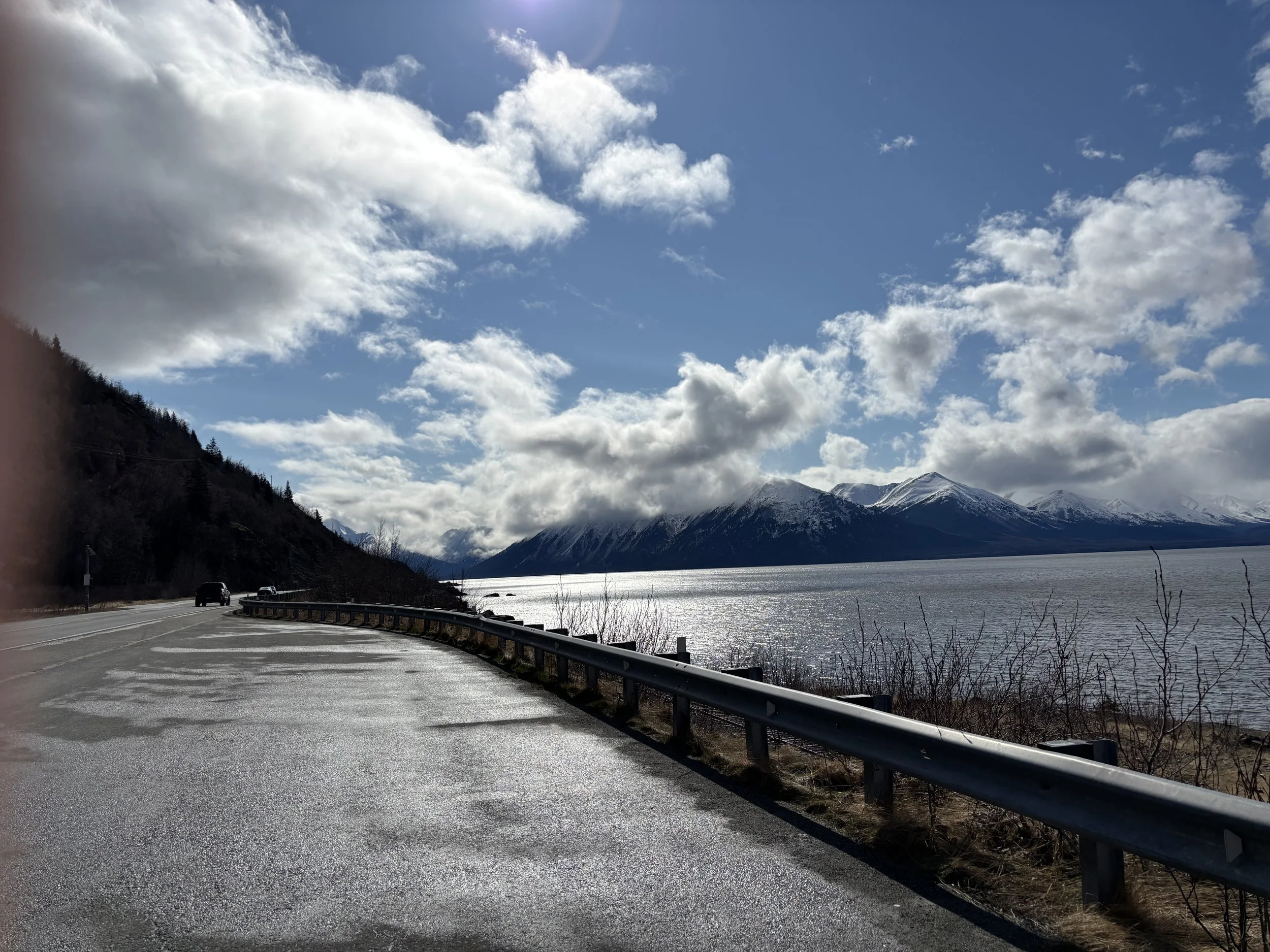 A scenic view of a highway running alongside a body of water with snow-capped mountains in the background. The sky is partly cloudy with patches of blue visible.