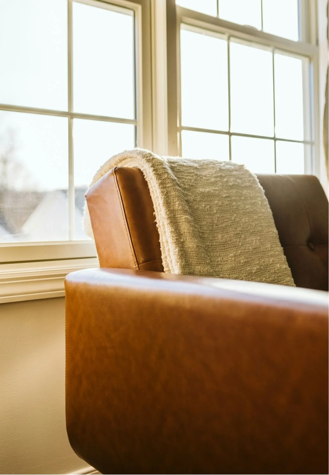 A leather armchair with a cozy blanket draped over the back, positioned in front of a large window letting in natural sunlight.