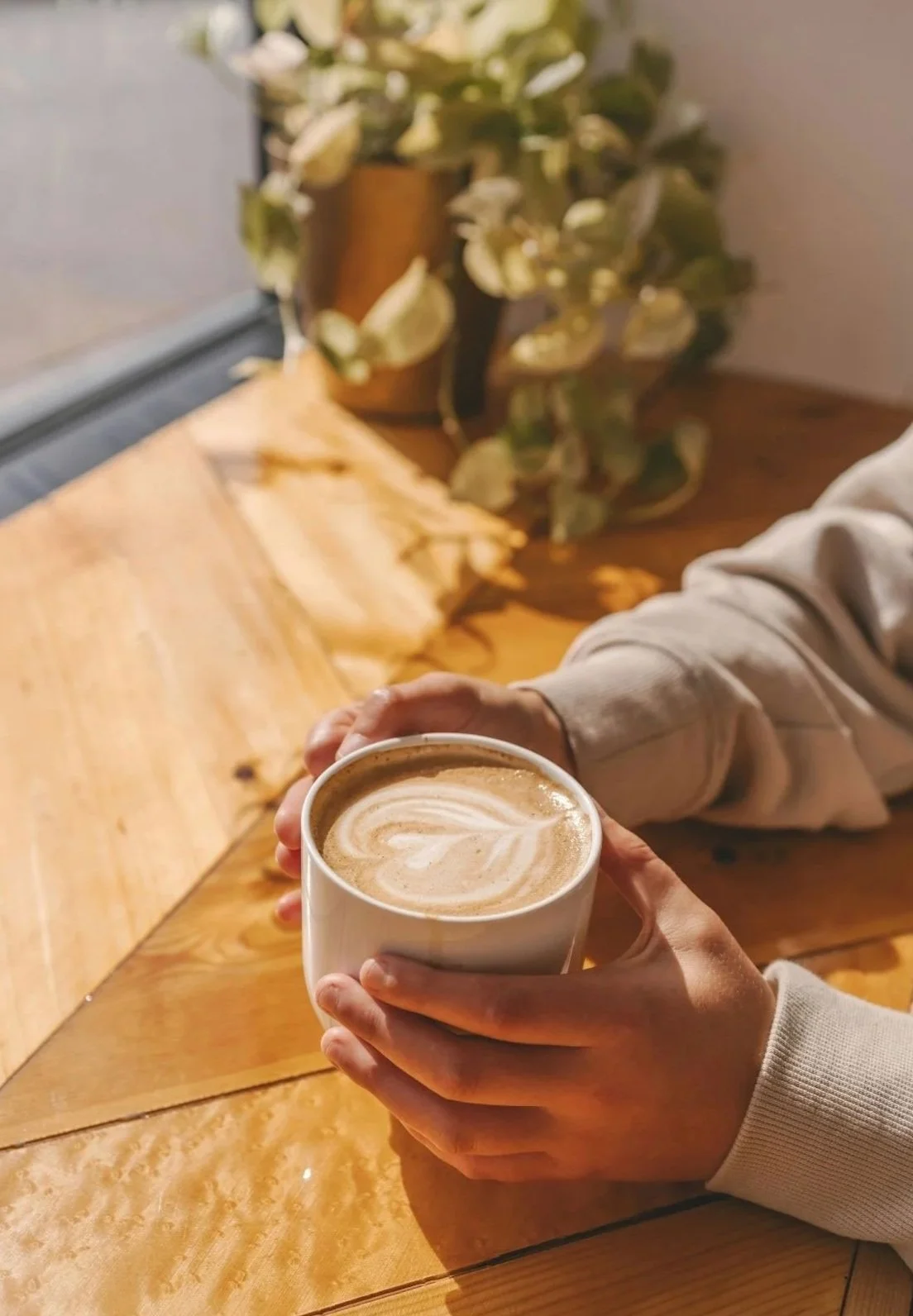 White person holding a cup of coffee in a cozy cafe setting, with a wooden table and a plant in the background.