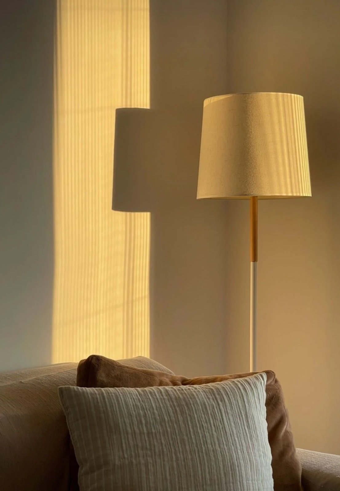 A beige floor lamp with a fabric shade casting a shadow on a wall behind a sofa with two pillows, one beige and one cream, in a warmly lit room.