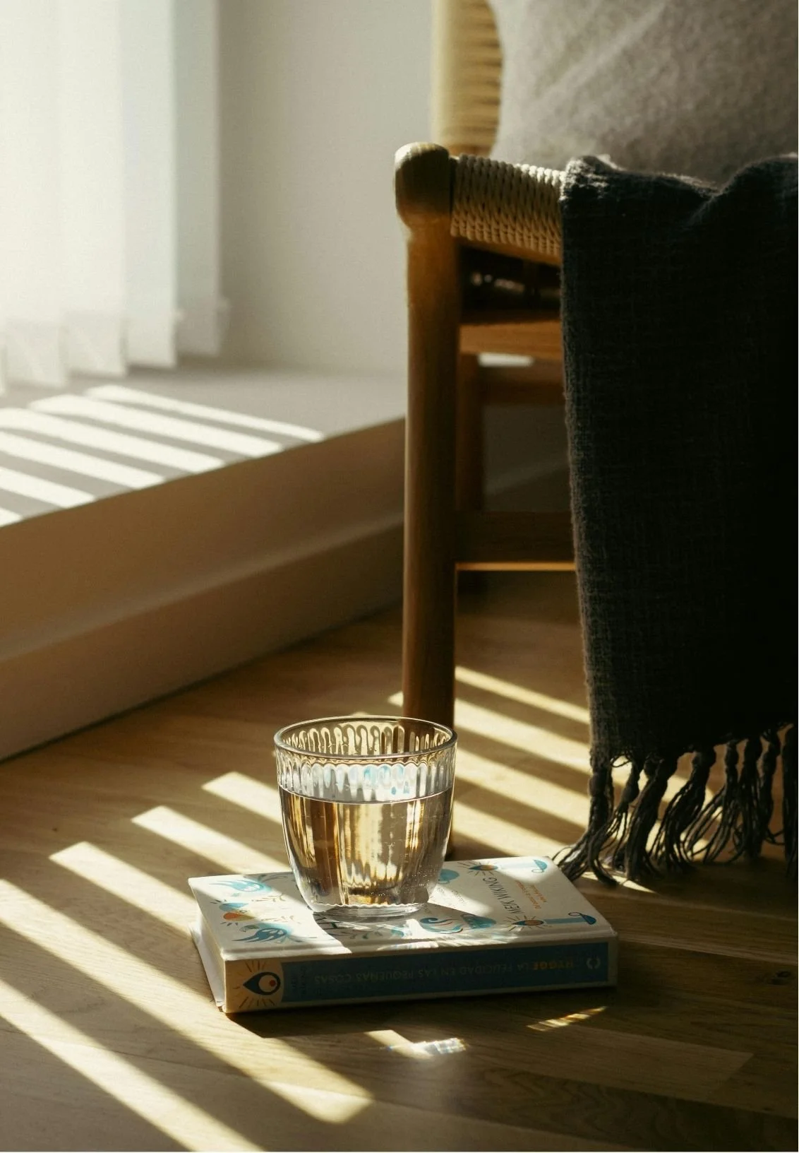 A glass of water placed on a book on a wooden floor, with sunlight casting shadows through a window. A chair with a black cloth draped over it is visible in the background.
