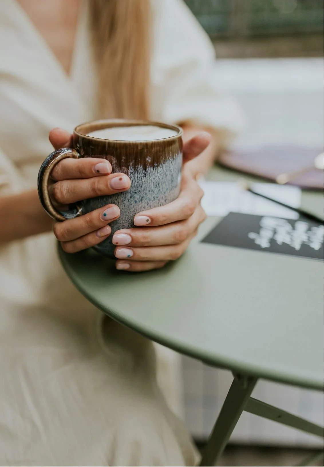 A white woman with painted nails holds a large, textured ceramic mug filled with a hot beverage. She is seated at a green round table with notebooks and papers nearby, outside near a window.