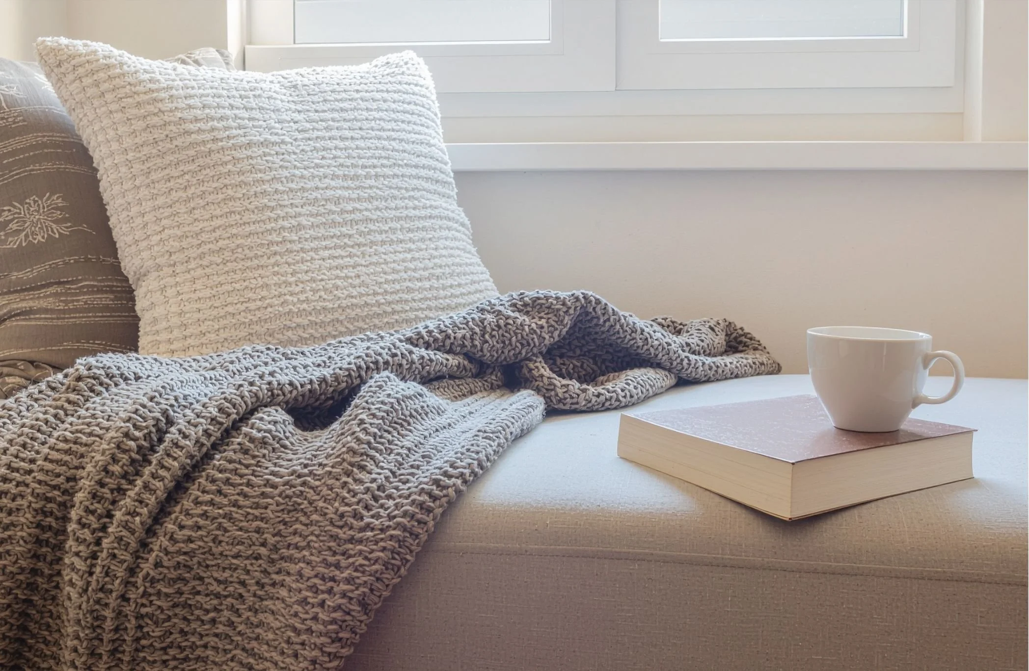 Close-up of a cozy, textured beige blanket and a large woven white pillow on a bed, with a white coffee mug resting on top of a large book on a light-colored bed surface near a window.