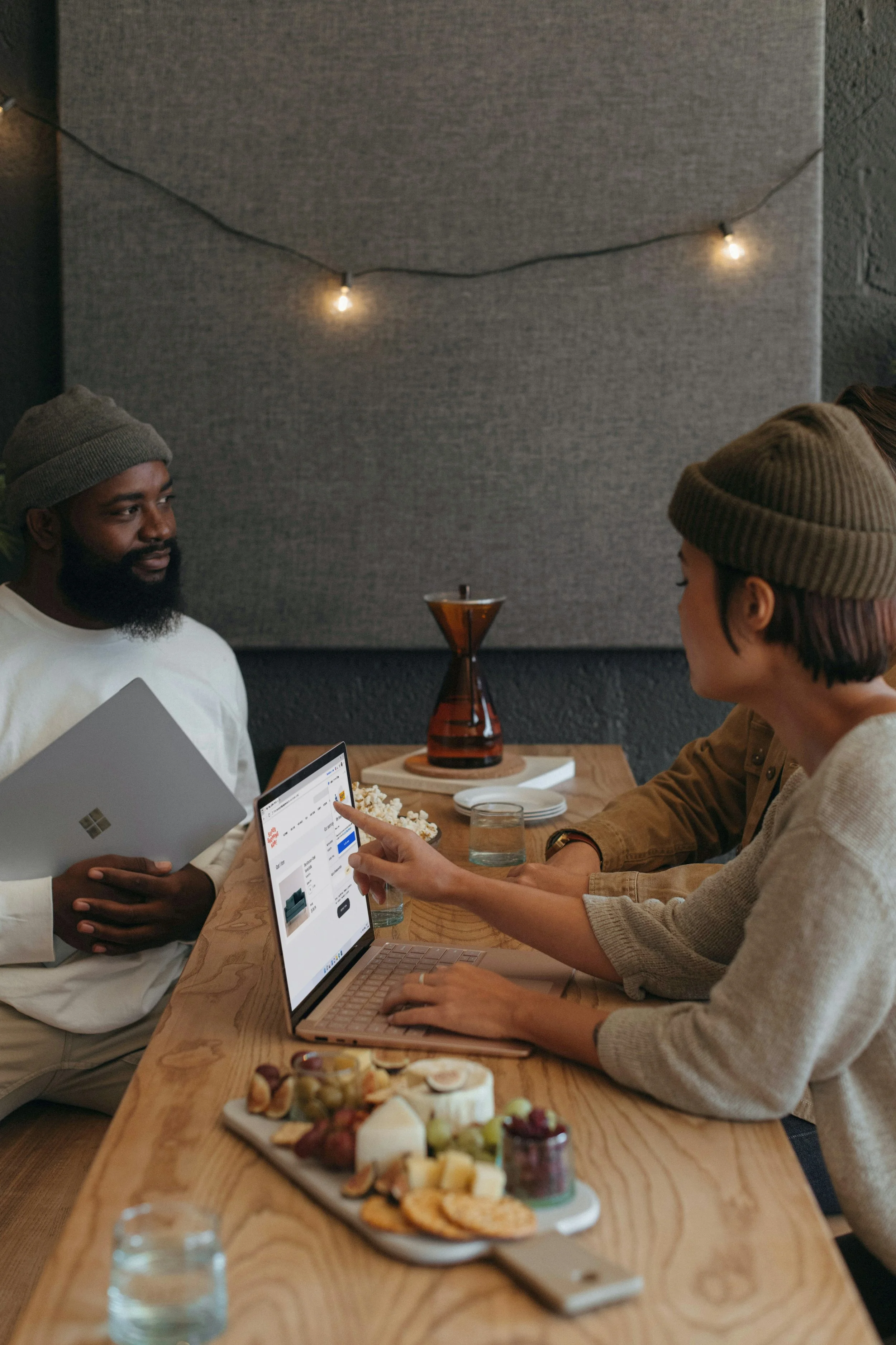 An African American person and an Asian American person sitting at a wooden table engaged in a conversation, with snacks and drinks on the table, and a gray wall with string lights in the background.