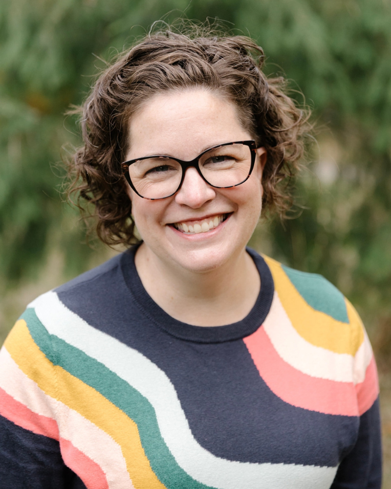 A smiling white woman with short curly brown hair and glasses, wearing a colorful striped sweater, outdoors with blurred green foliage in the background.