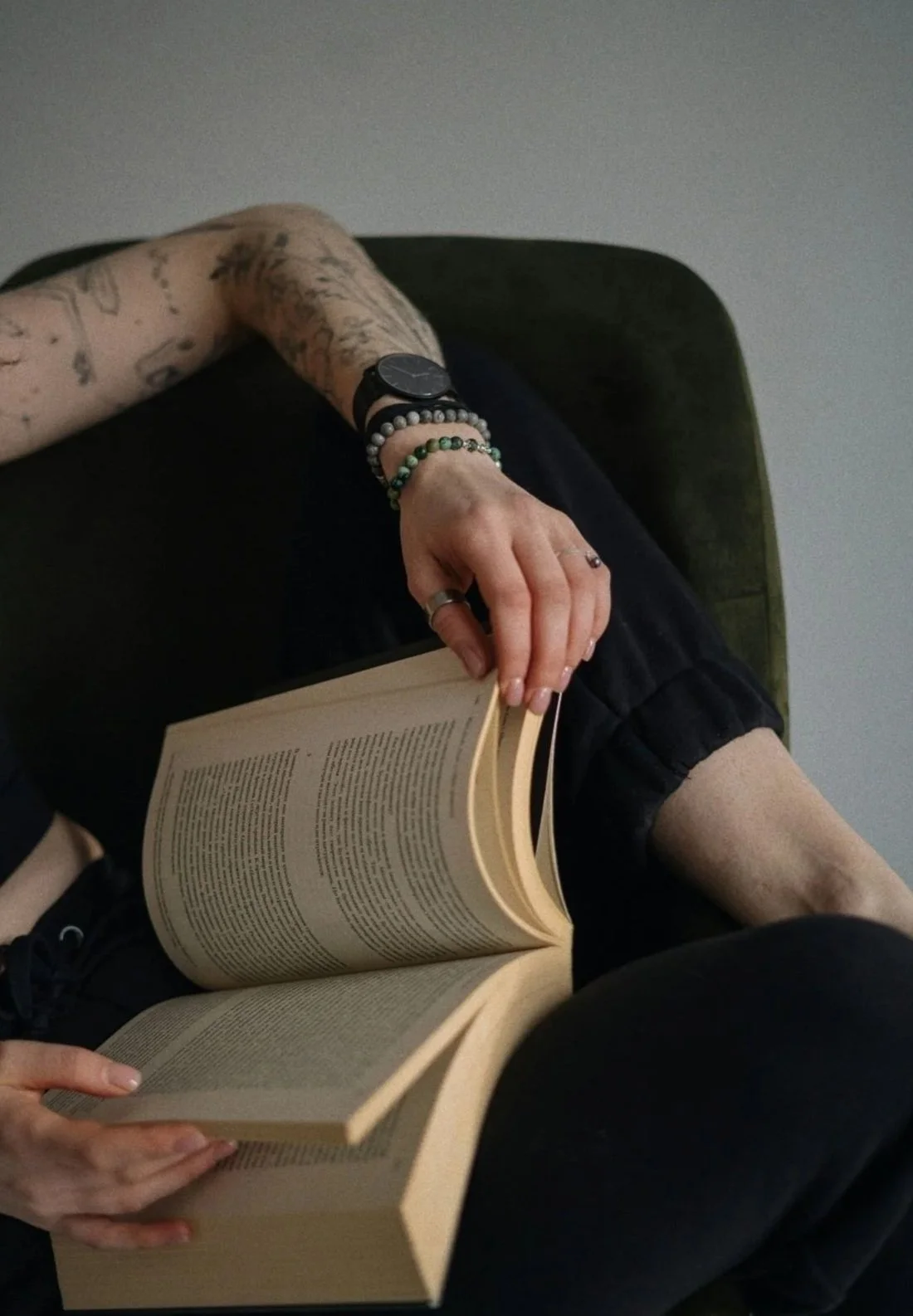 A person sitting on a green chair or sofa, reading a book. They have tattoos on their arm, are wearing a watch and bead bracelets, and have rings on their fingers.