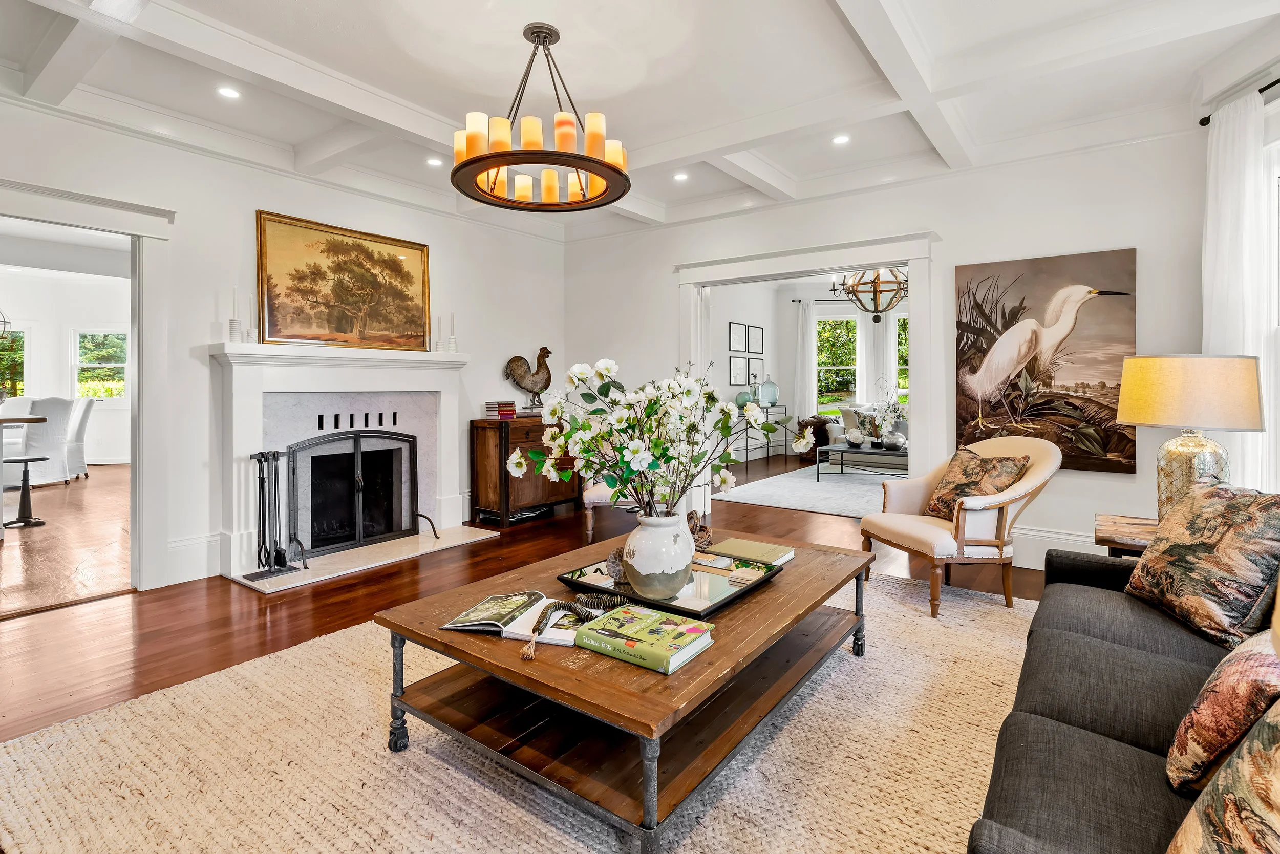 Living room with white walls, hardwood floors, a fireplace, floral arrangements on a coffee table, and wall art including a painting of a tree and a photograph of a heron.