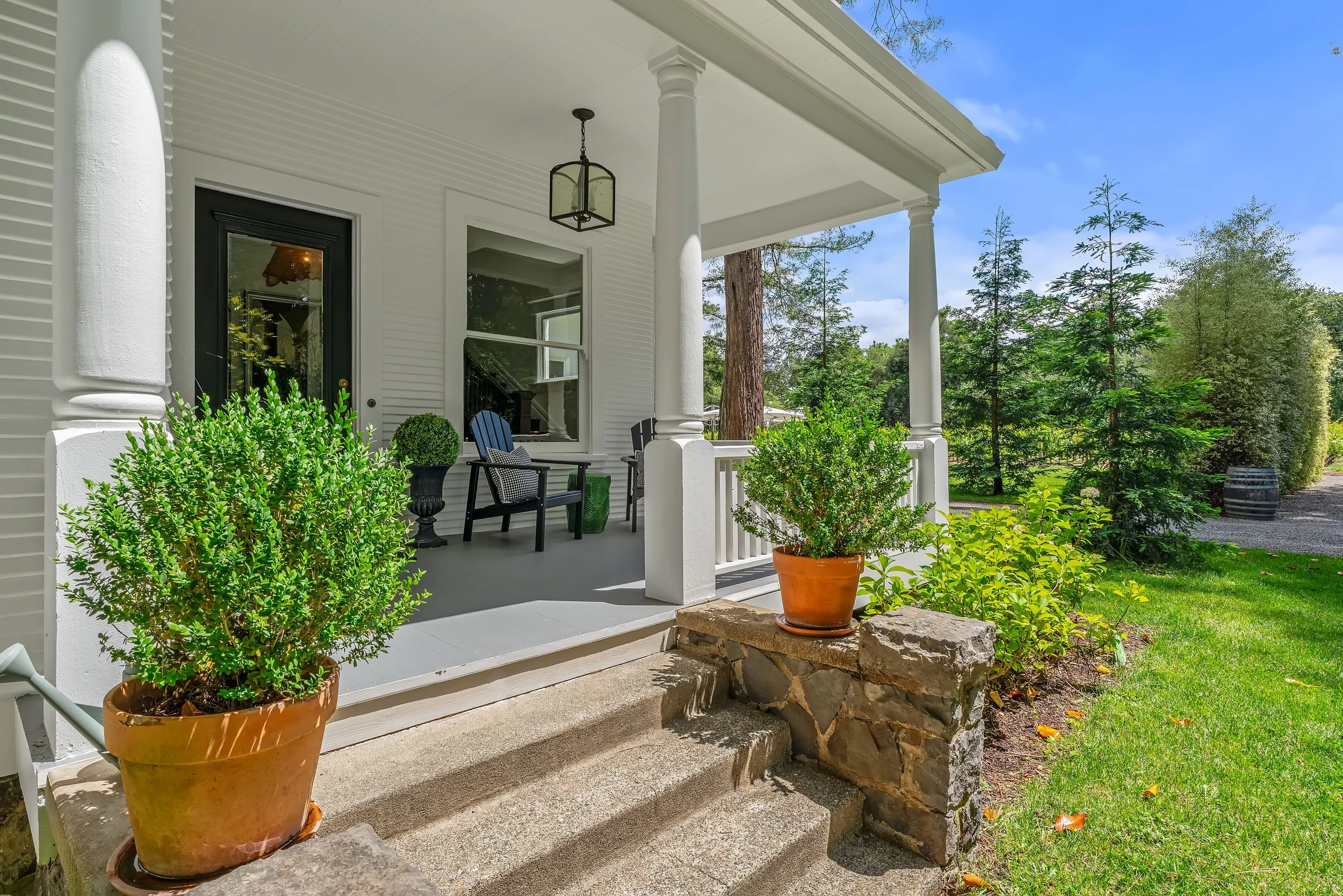 Front porch of a white house with black door and large window, furnished with black chairs and potted plants, surrounded by greenery and a blue sky.