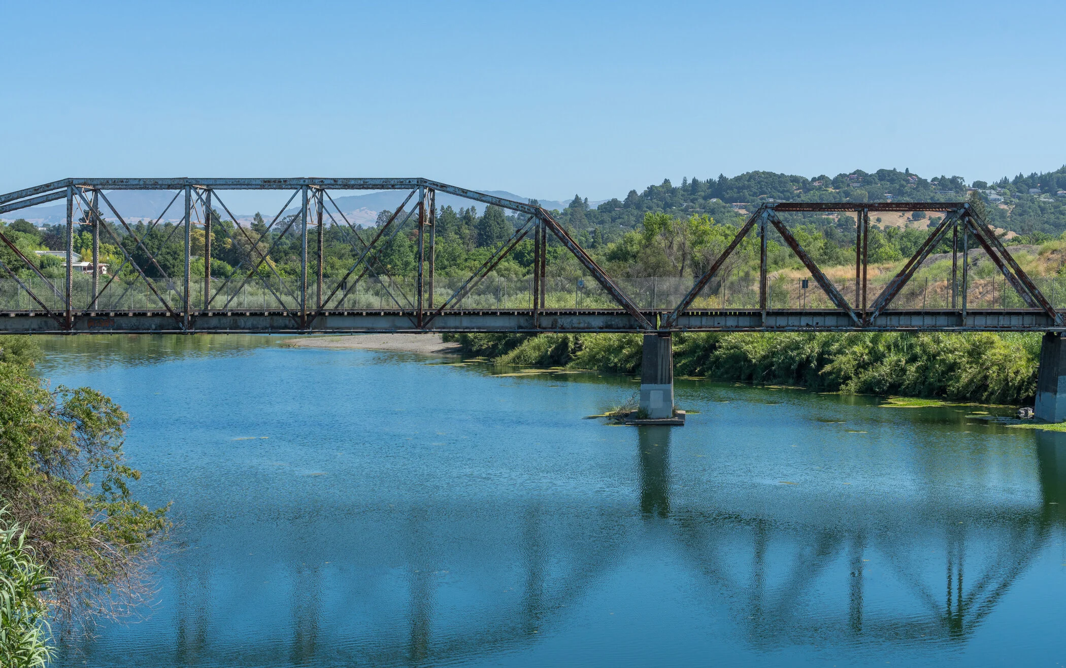An old rusted steel bridge spanning a calm river with green trees along the riverbanks and rolling hills with houses in the background under a clear blue sky.