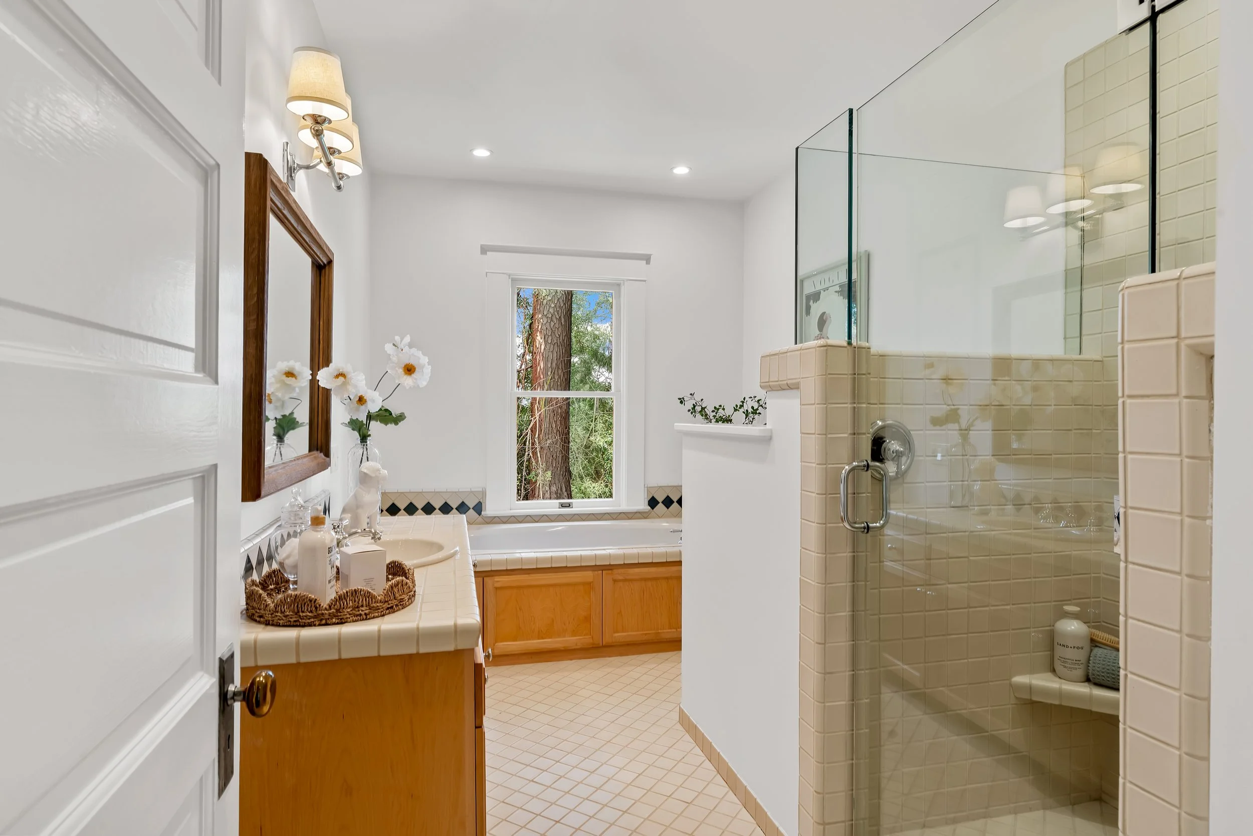 Bathroom with white walls, a window showing trees outside, a vanity with a mirror, a bathtub, and a shower stall with beige tiles.
