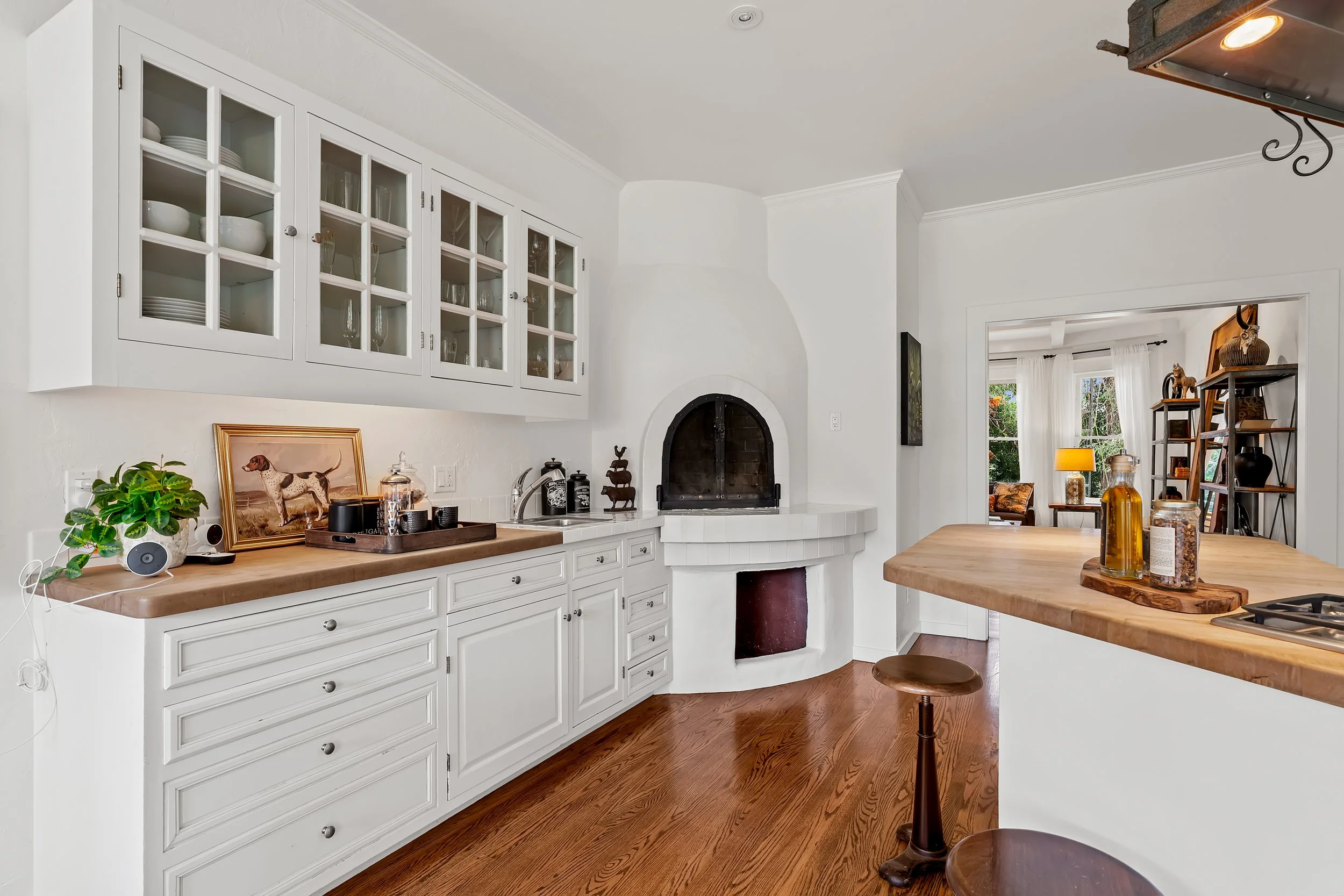 A bright kitchen with white cabinetry, a wooden kitchen island, a fireplace, and a doorway leading to a living room with large windows and white curtains.