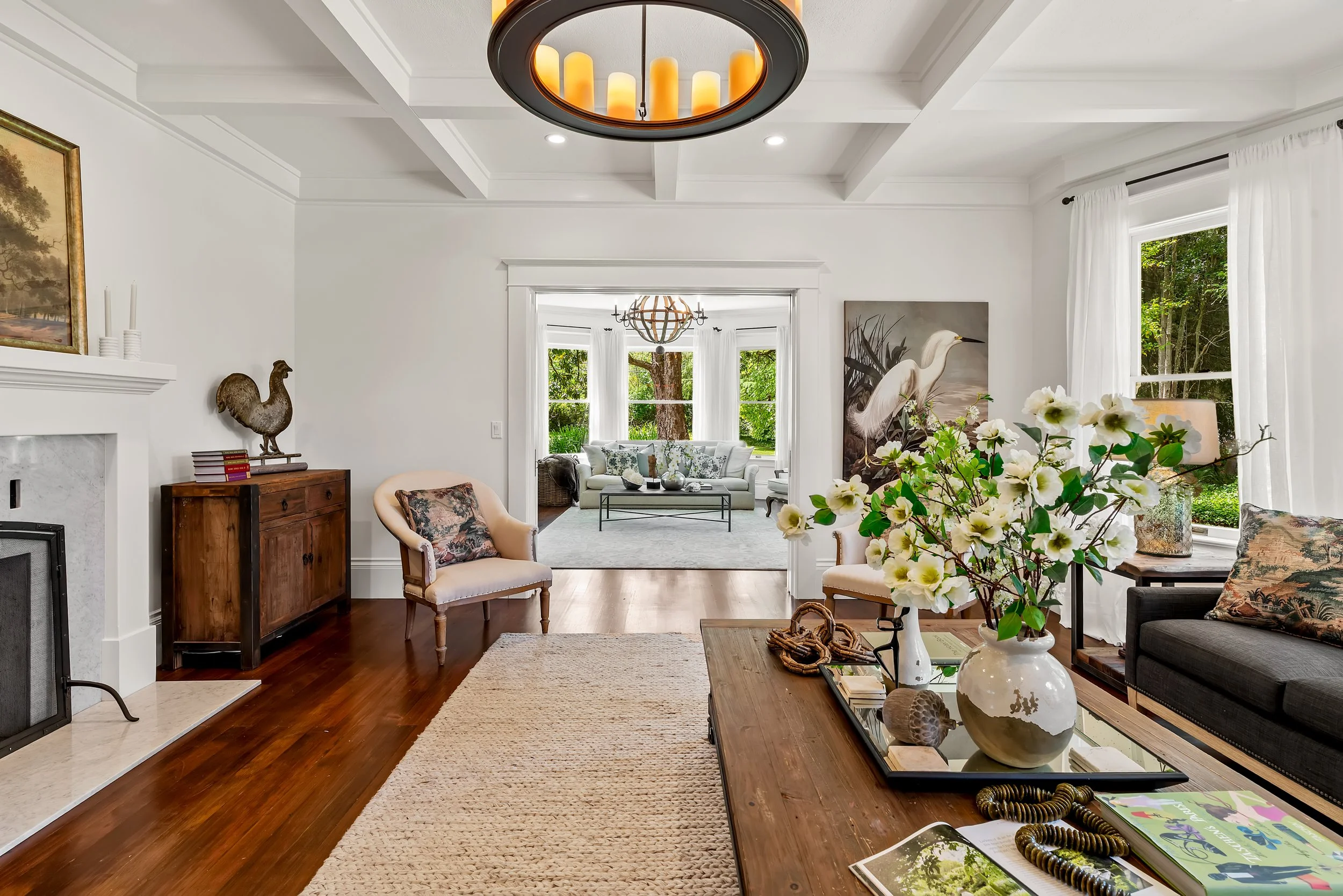 Bright living room with white walls and large windows, decorated with artwork, plants, and furniture, including chairs and a coffee table, illuminated by chandeliers and natural light.