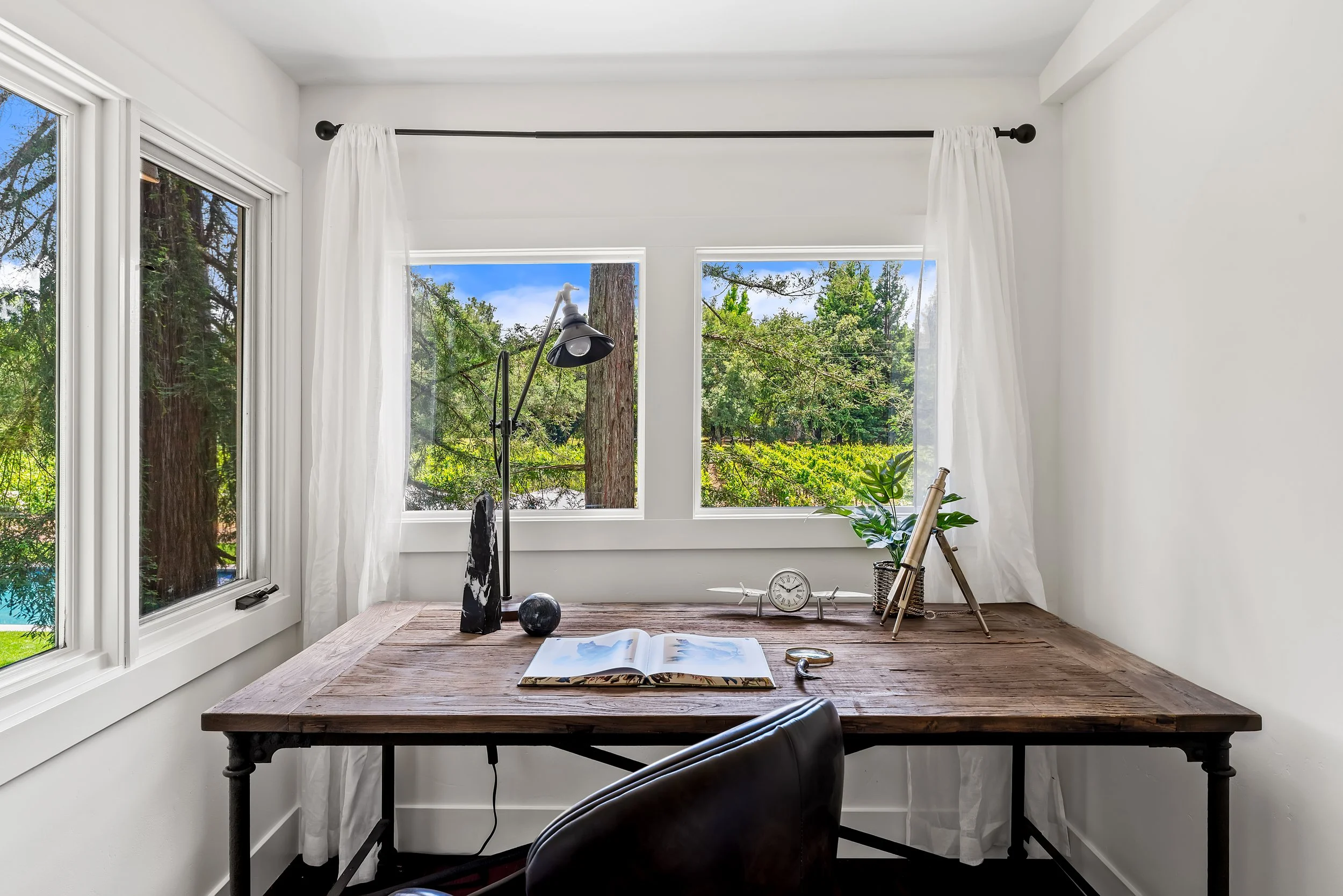 A minimalist home office with a wooden desk, white walls, large windows showing green trees outside, and various decor items including a black lamp, a book, and a small clock.