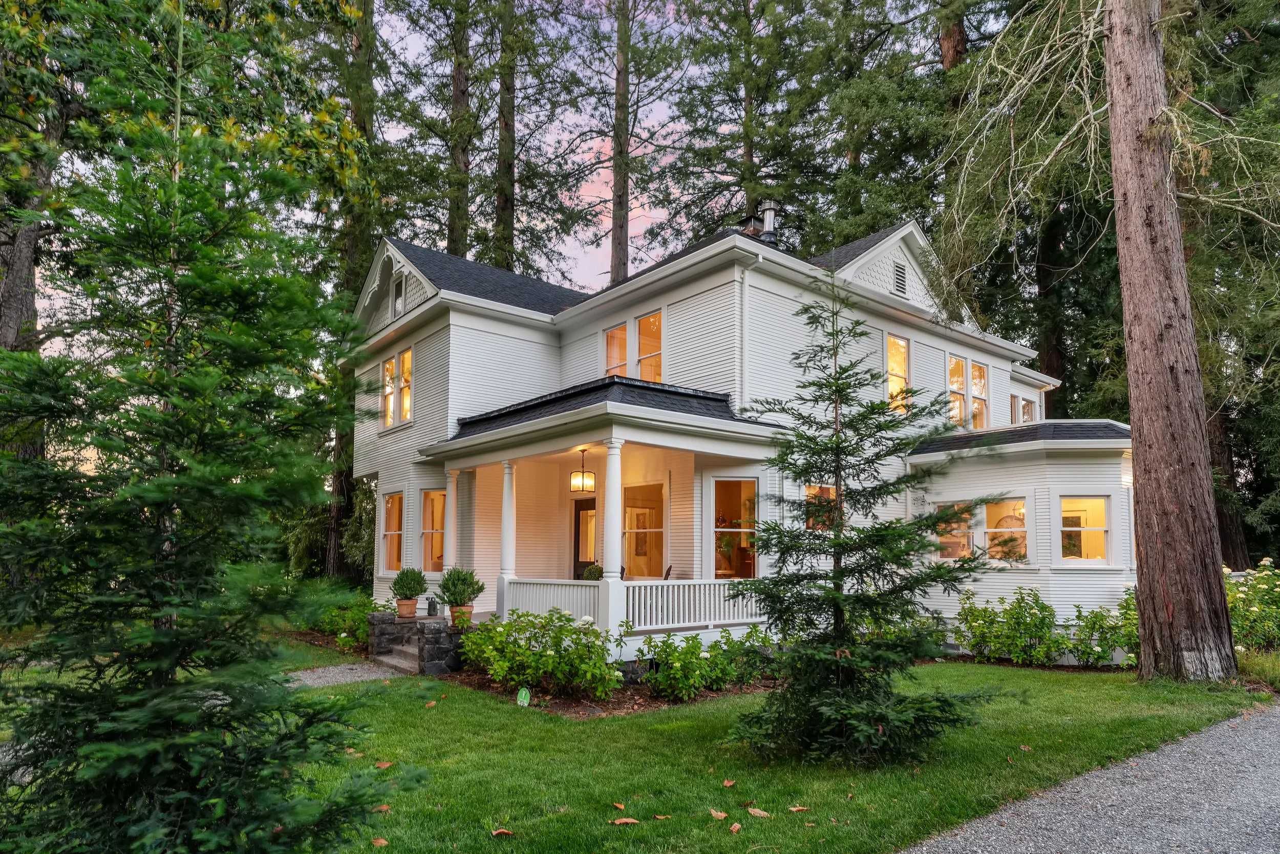A two-story white house surrounded by tall trees and lush green lawn, with warm light glowing from the windows during dusk.