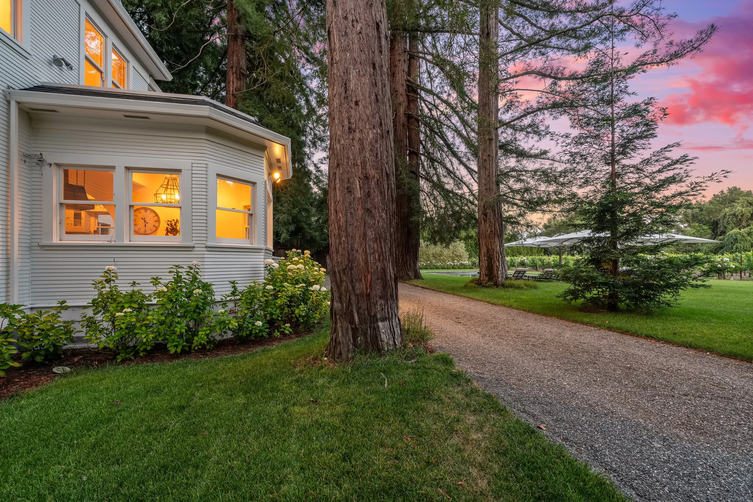 A white house with large windows glowing with warm light, surrounded by green bushes and tall trees, during an evening with a colorful pink and purple sunset sky.