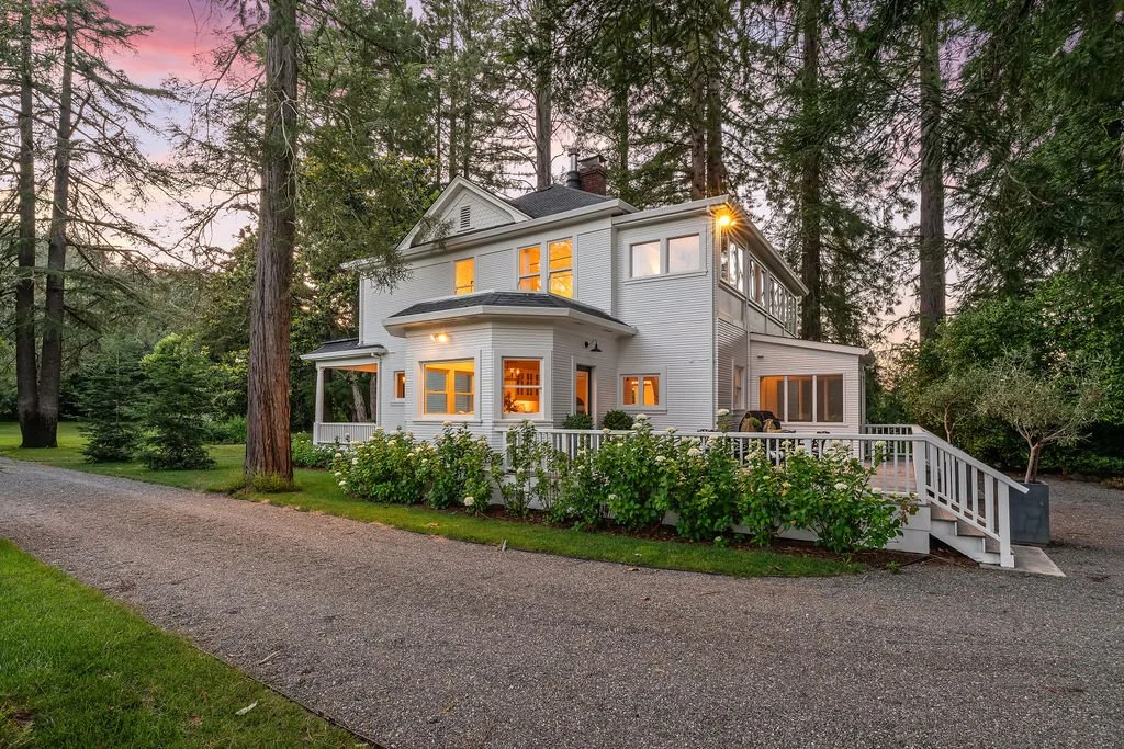 A large, white, two-story house with multiple windows, a porch, and a deck, surrounded by tall trees and greenery at dusk.