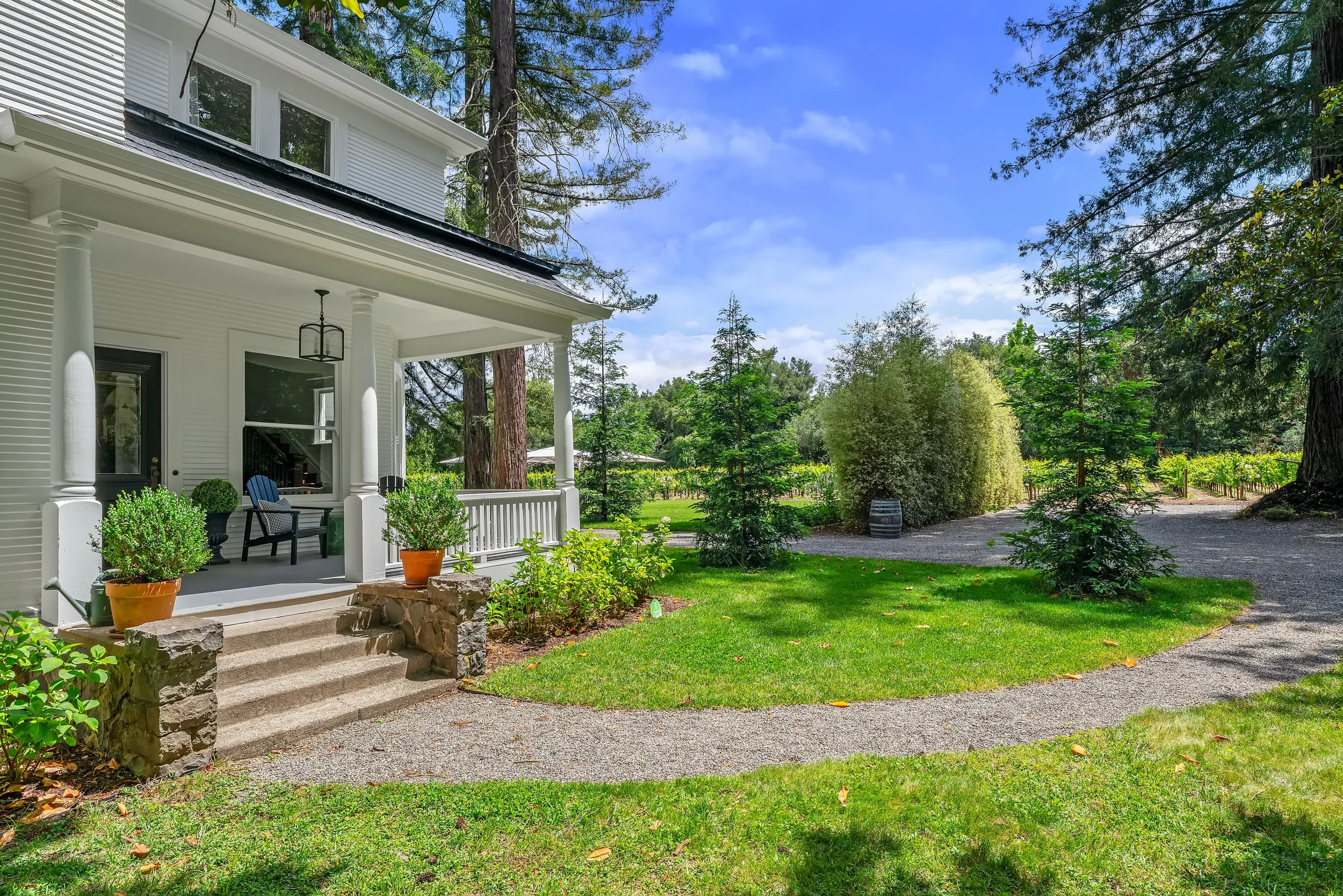 A white house with a porch, potted plants, and a hanging lantern, surrounded by trees, with a gravel driveway and green lawn.