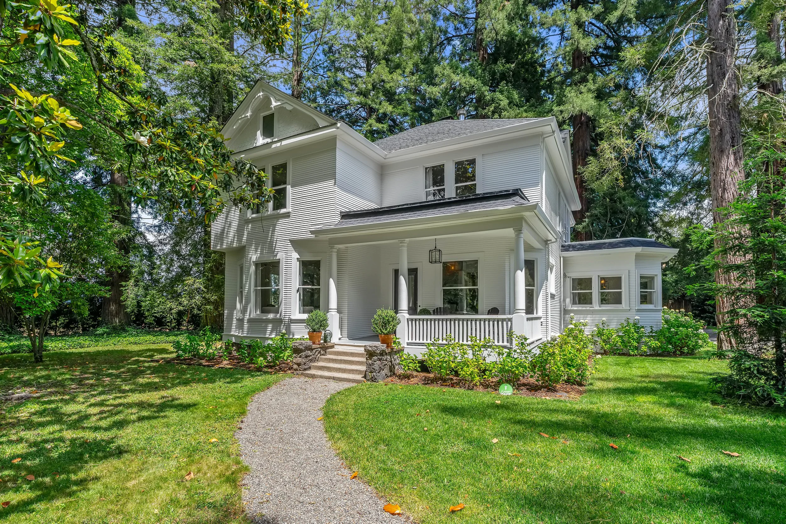 White, two-story house with a front porch, surrounded by lush green trees and grass, with a gravel pathway leading up to the stairs.