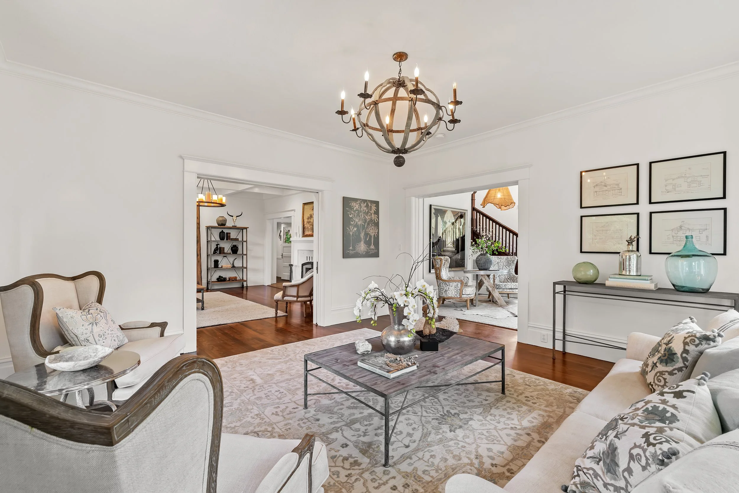 A spacious living room with white walls and wooden floors. There are two cream-colored sofas with patterned cushions, a wooden coffee table with decorative items and a stack of books, and a side table with a large glass vase. Artwork decorates the wa