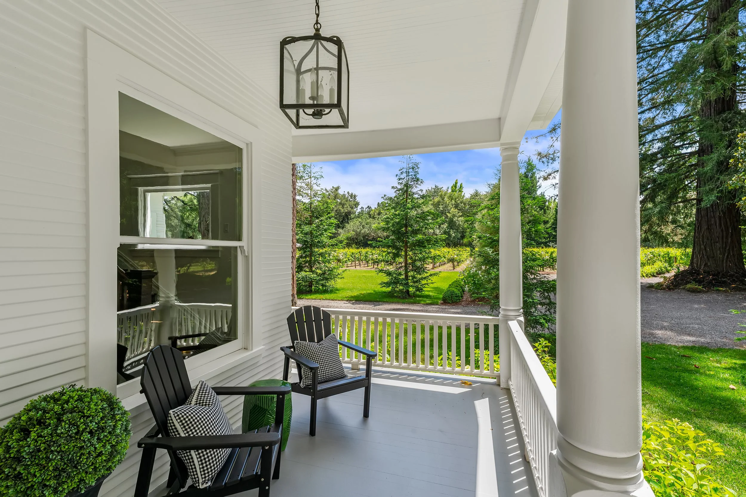 A porch with black chairs and checkered cushions, a small green plant, and a large round shrub, surrounded by white columns and railings, overlooking a lush garden with trees and a vineyard under a blue sky.