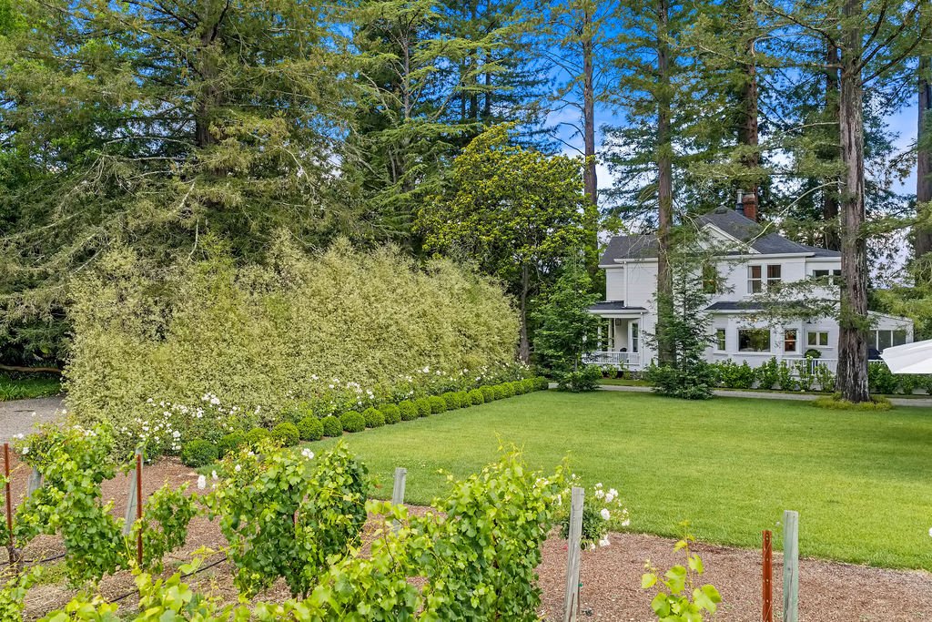 A large white house with multiple stories, surrounded by tall trees and a well-maintained lawn. There are landscaped bushes and a garden in the foreground.
